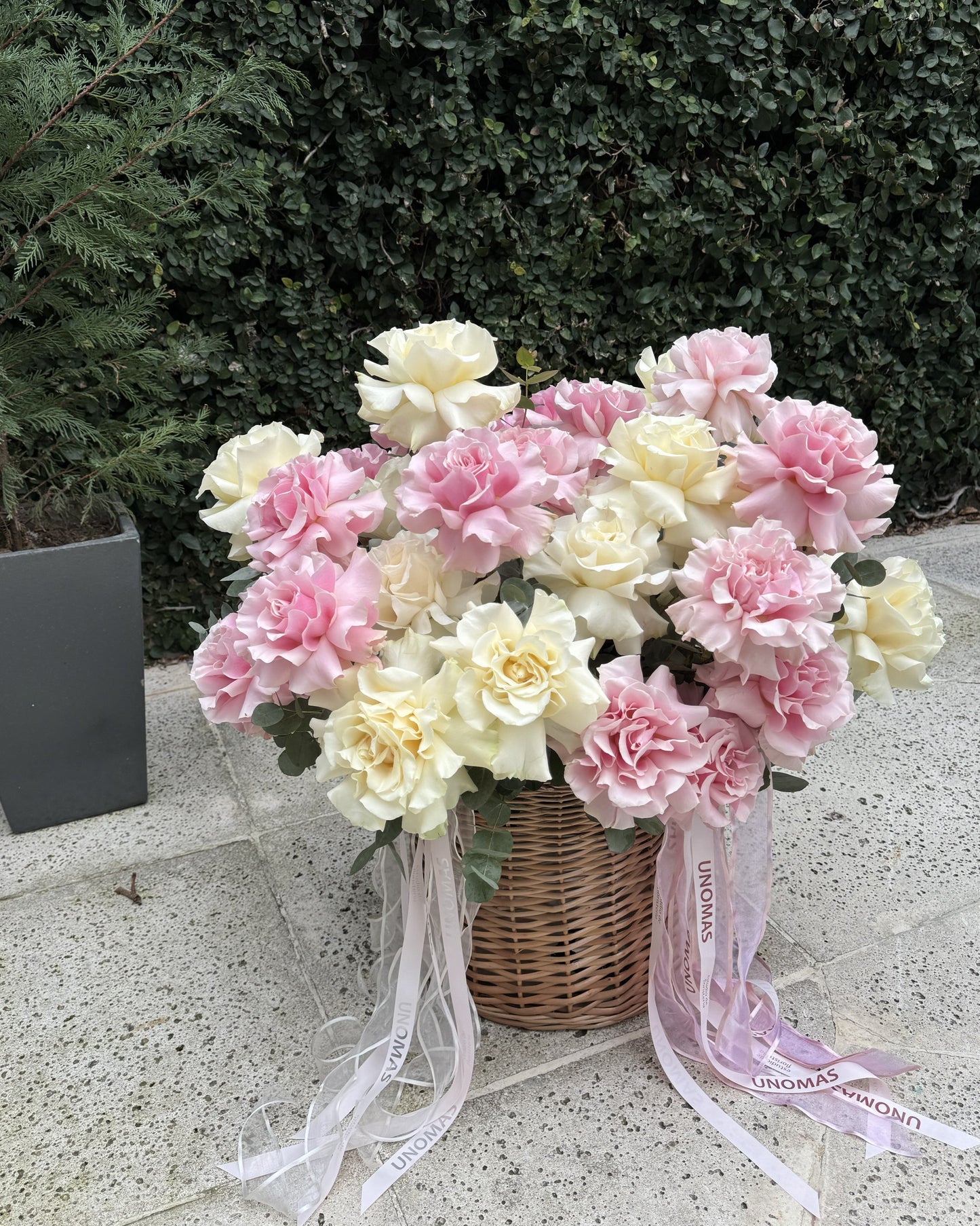 A bouquet of roses in shades of pink and cream, arranged in a wicker basket with white and pink ribbons, set on a stone patio with a green hedge background.