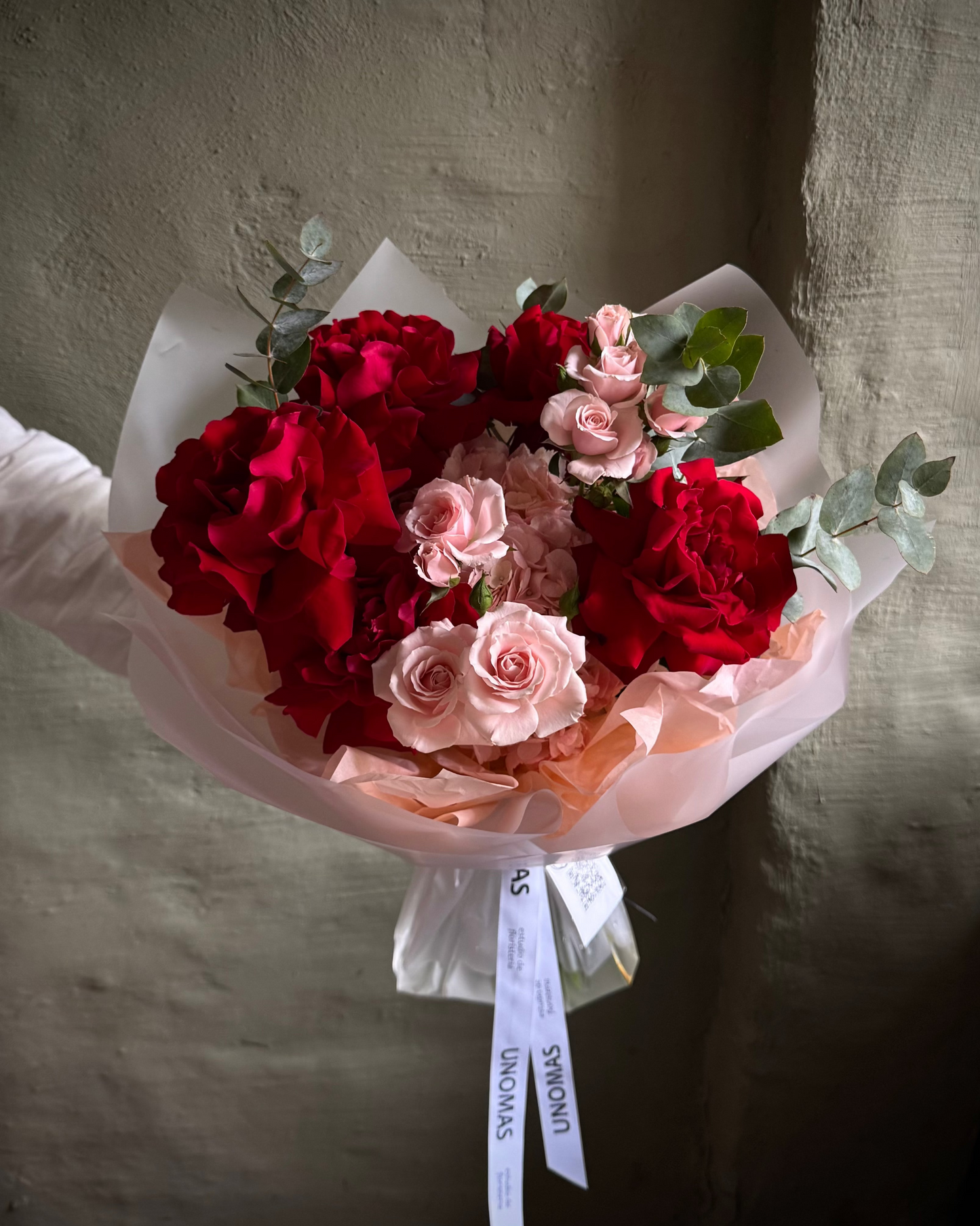 Bouquet of red and pink flowers wrapped in paper with a visible brand label against a textured wall.