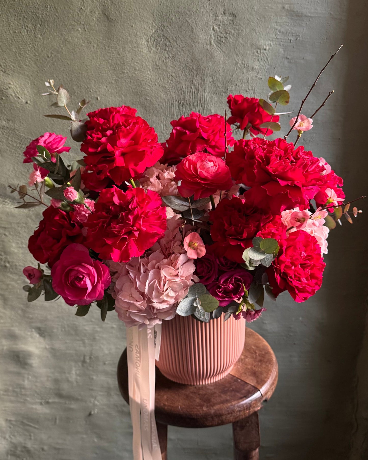 Bouquet of red and pink flowers in a pink vase on a wooden stool against a gray wall.
