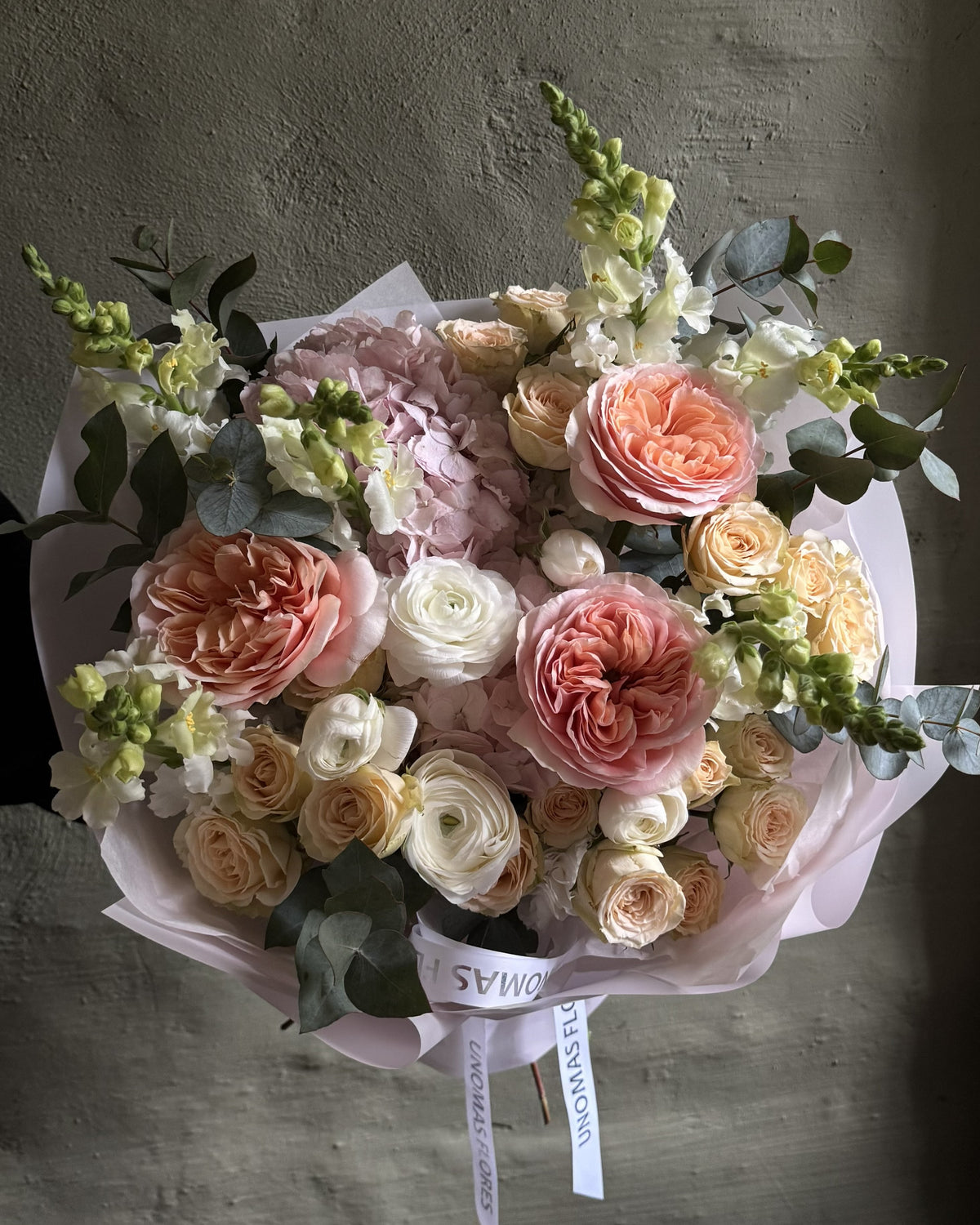 Ramo de rosas durazno y crema, ranúnculos y hortensias rosadas con eucalipto, envuelto en papel rosa claro.