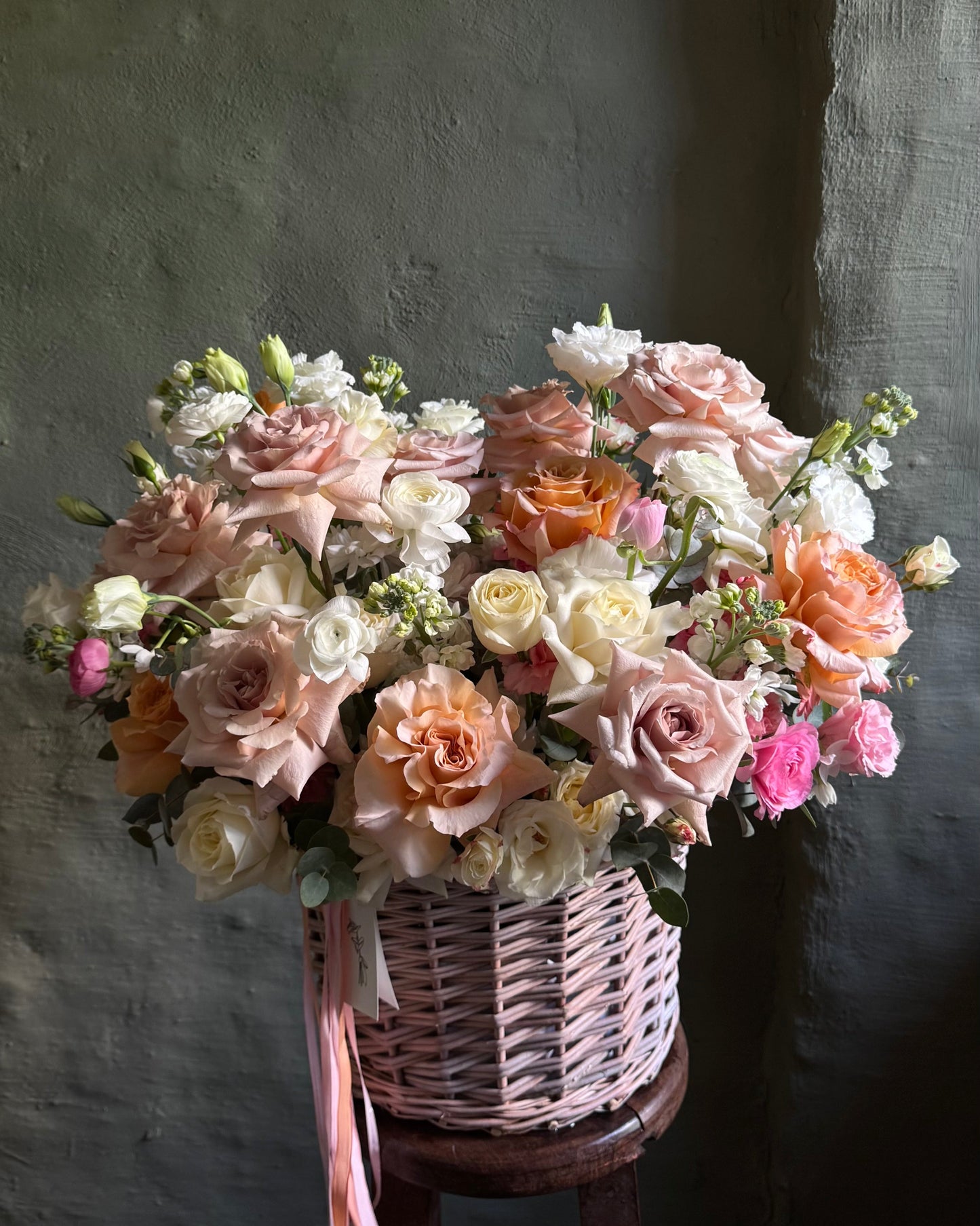 Flower arrangement in a wicker basket of peach roses of the Shimmer variety, local roses, bush roses, soft pink ranunculus and matthiola