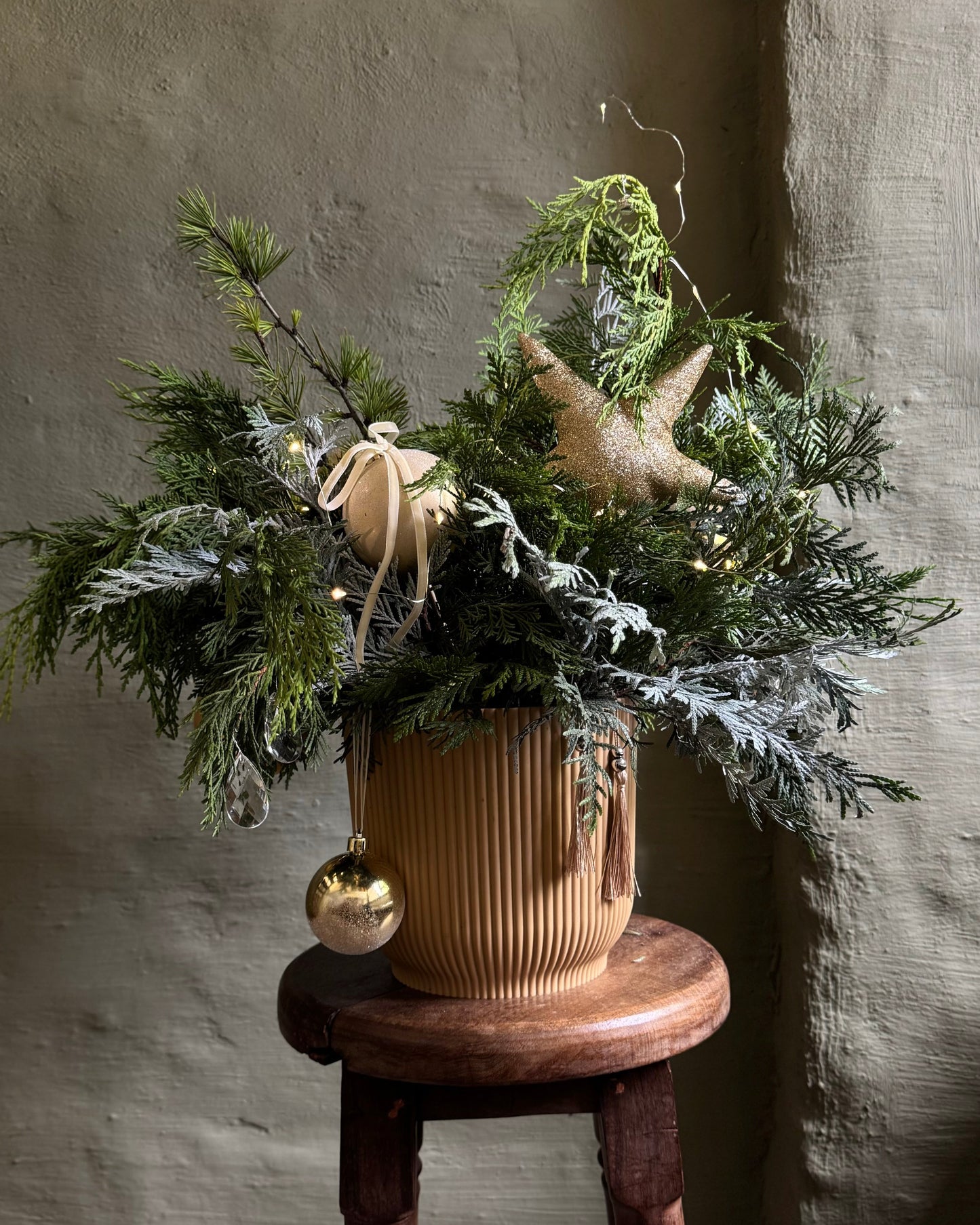 Decorative plant arrangement with ornaments on a wooden stool against a textured wall.