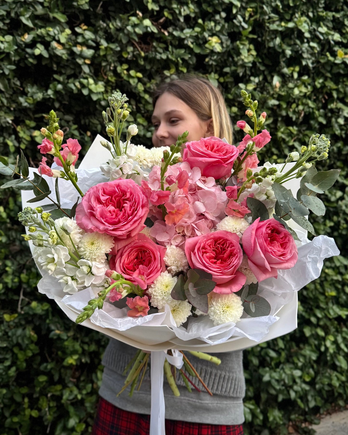 Person holding a large bouquet of pink and white flowers against a green hedge.