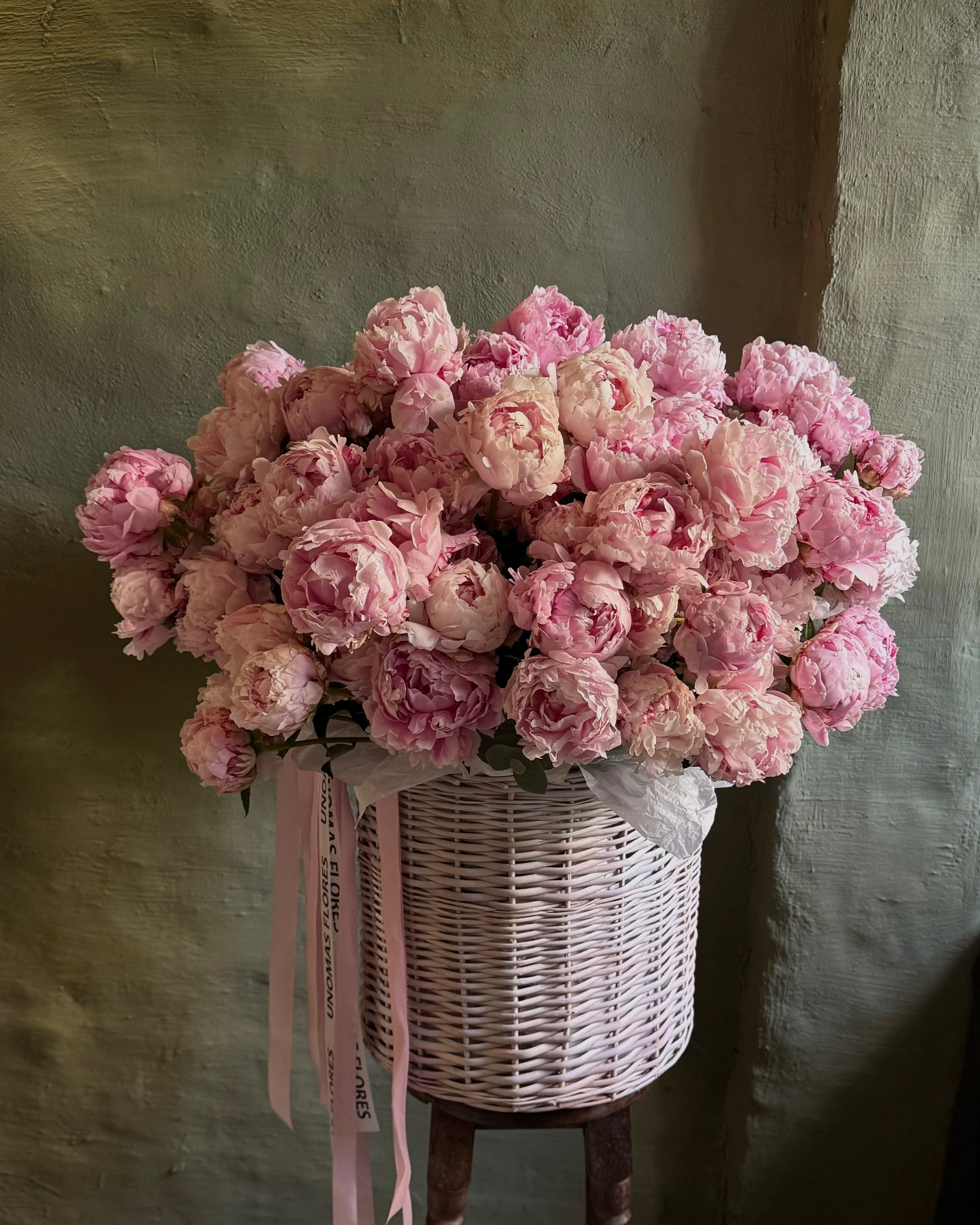 Bouquet of pink flowers in a wicker basket on a wooden stool against a textured wall.