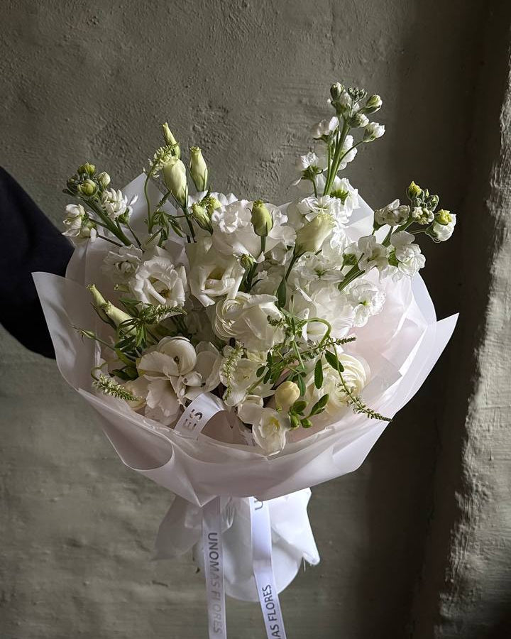 Ramo de flores blancas de estación con alhelíes, conejitos y follaje, envuelto en papel blanco y atado con una cinta.