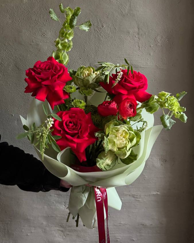 Ramo de rosas rojas y verdes con eucalipto, envuelto en papel verde claro y atado con una cinta bordó.