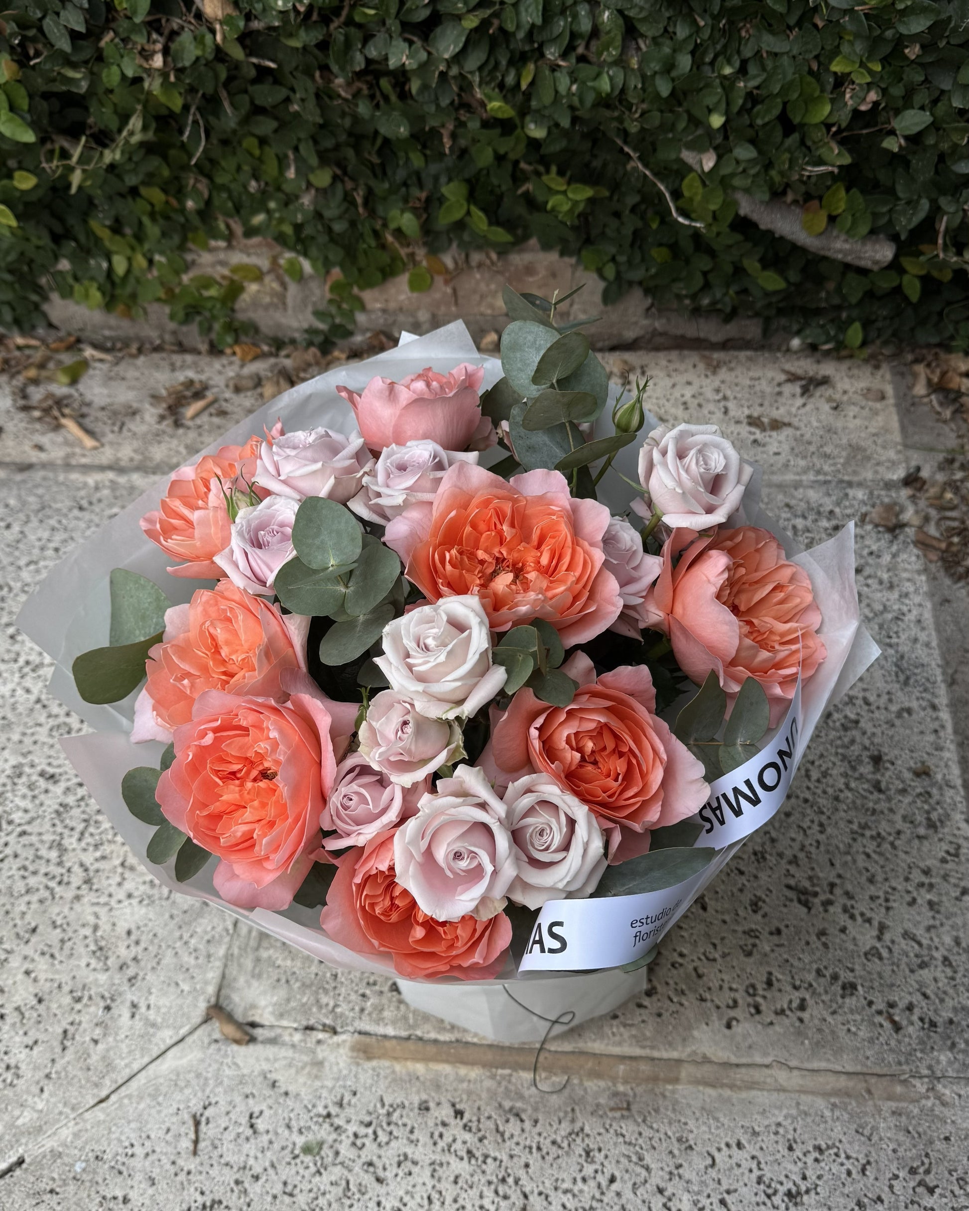 A bouquet of mixed flowers, including coral roses, light pink roses with green eucalyptus leaves, wrapped in white tissue paper and a white ribbon, placed on a stone surface with green foliage in the background.