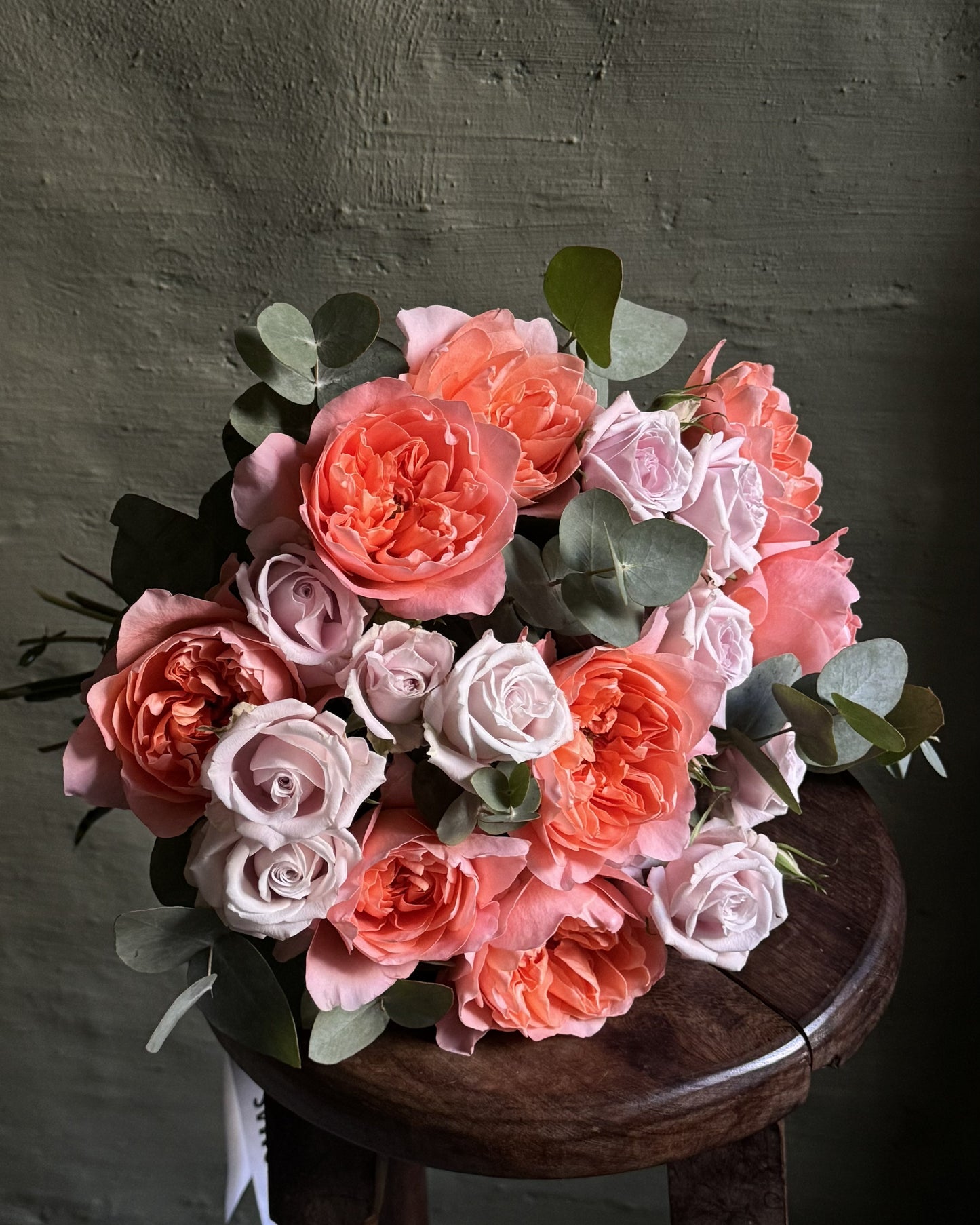 A bouquet of mixed roses in shades of coral, peach, pink, and pale lavender, accented with green eucalyptus leaves, displayed on a dark wooden stool against a textured dark gray wall.