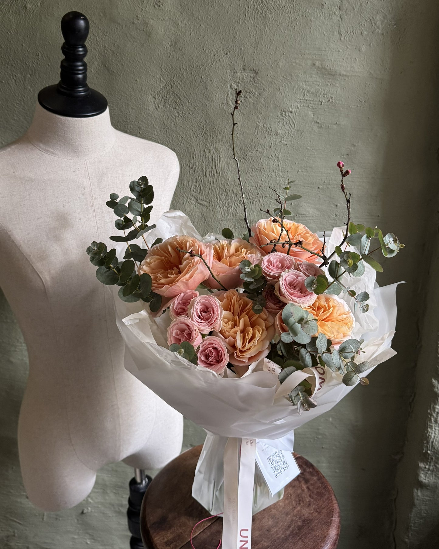 A floral bouquet with peach and pink roses, eucalyptus leaves, and delicate stems, wrapped in white tissue paper and tied with a white ribbon, displayed next to a cream fabric mannequin torso on a wooden stool against a textured sage green wall.
