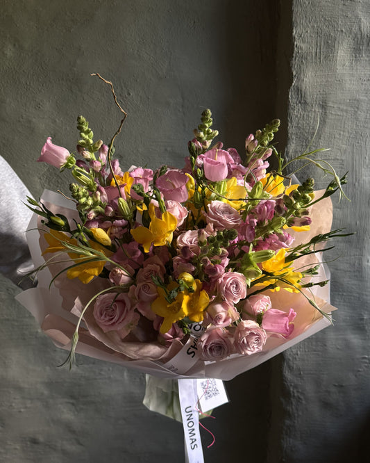 A hand-held bouquet of pink and yellow flowers wrapped in pale pink paper, with a white ribbon tag printed with the brand name 'UNOMAS', set against a textured gray wall.