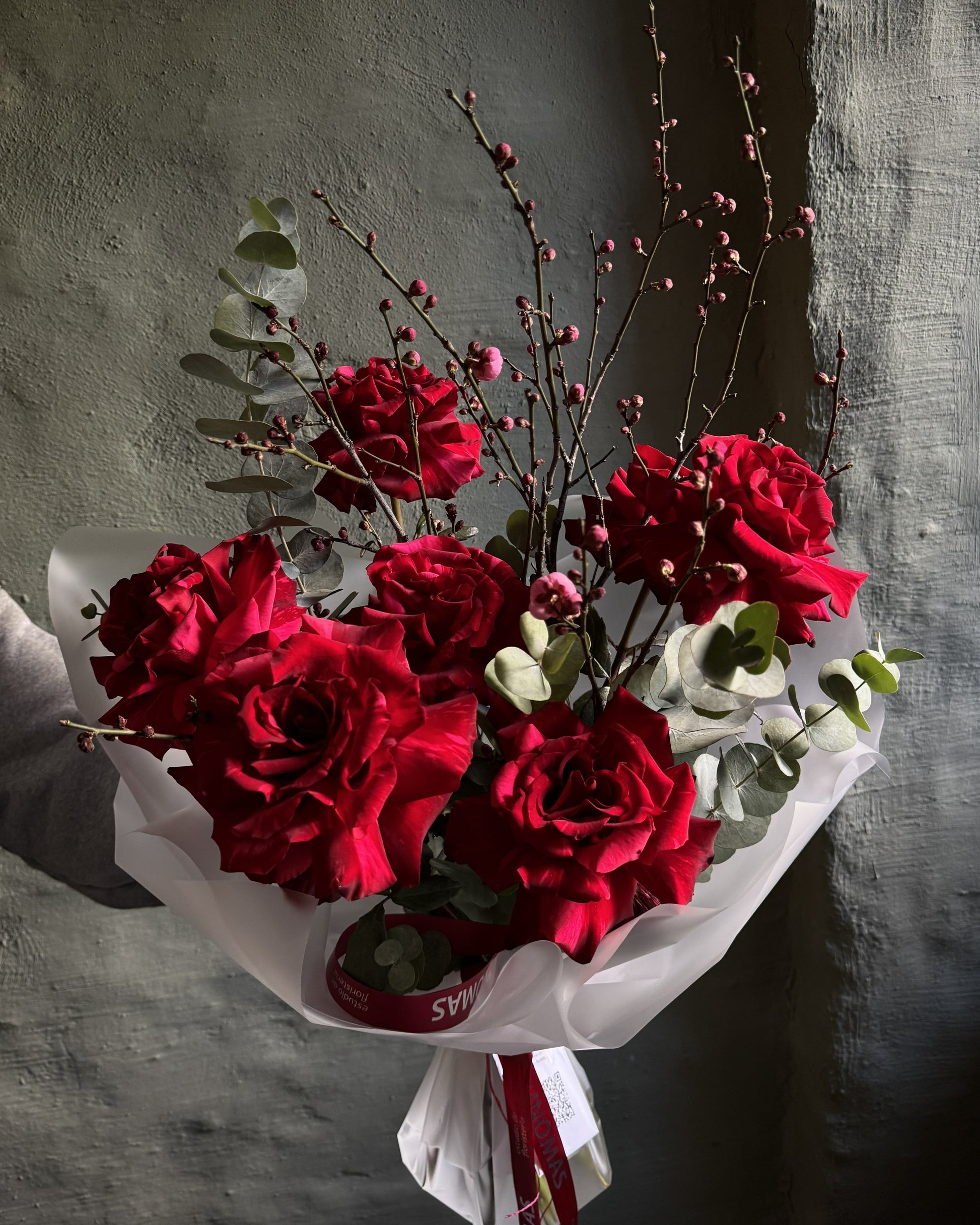 A premium bouquet of deep red roses with green eucalyptus leaves and delicate pinkish-brown twig branches, wrapped in white tissue paper and tied with a red ribbon