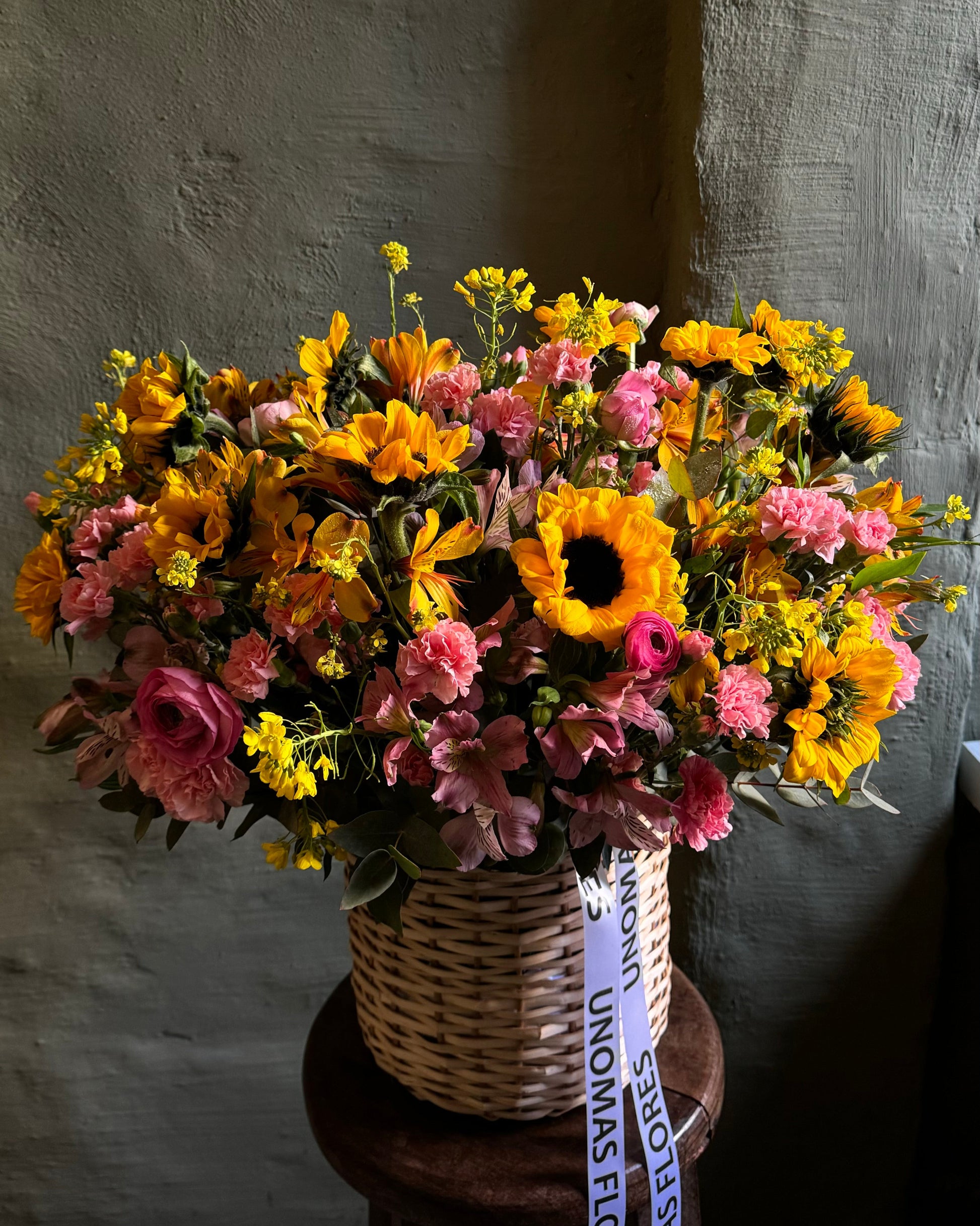 Bouquet of flowers in a woven basket with a visible brand label against a textured wall.