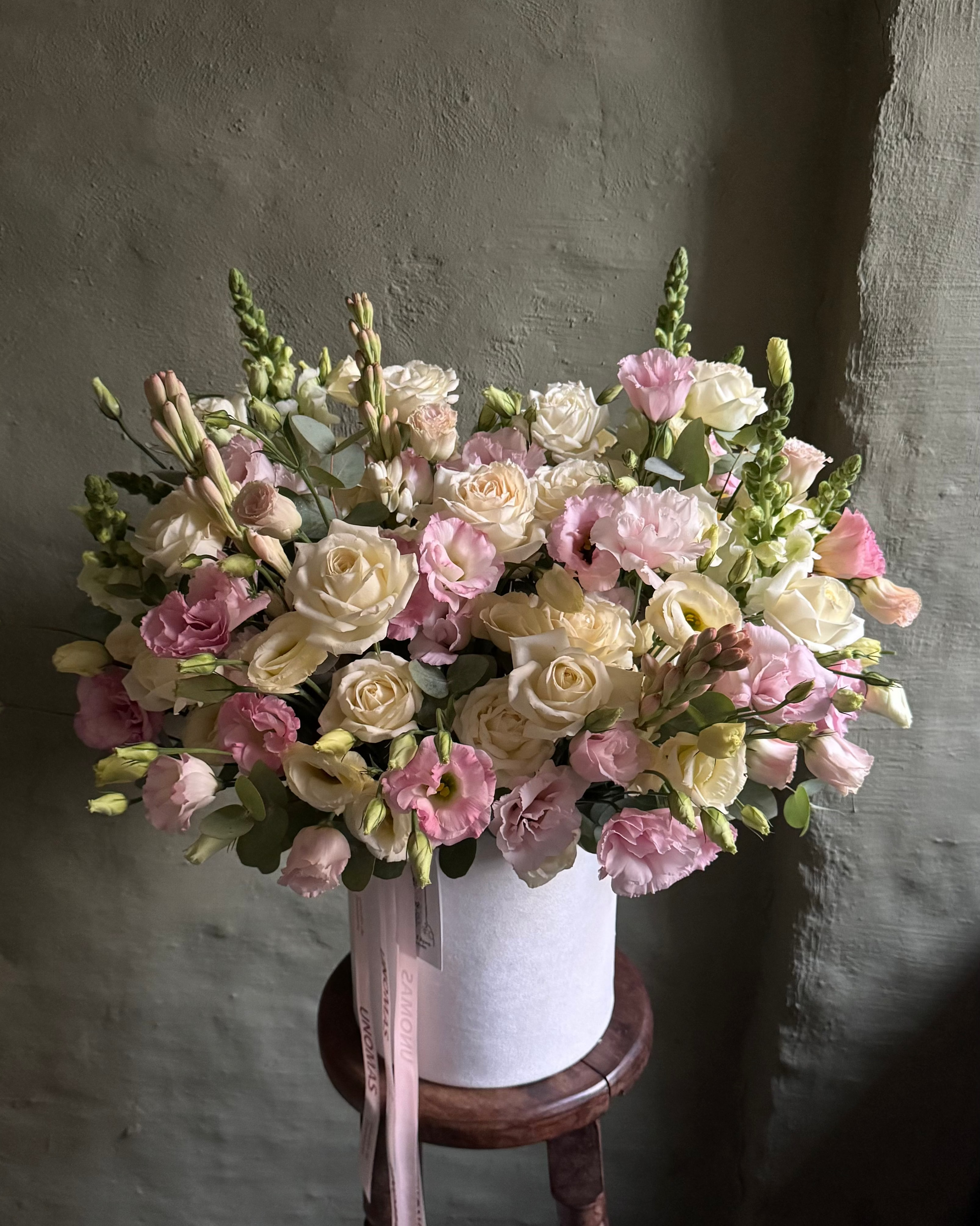 Bouquet of flowers in a white cachepot on a wooden stool against a gray wall.