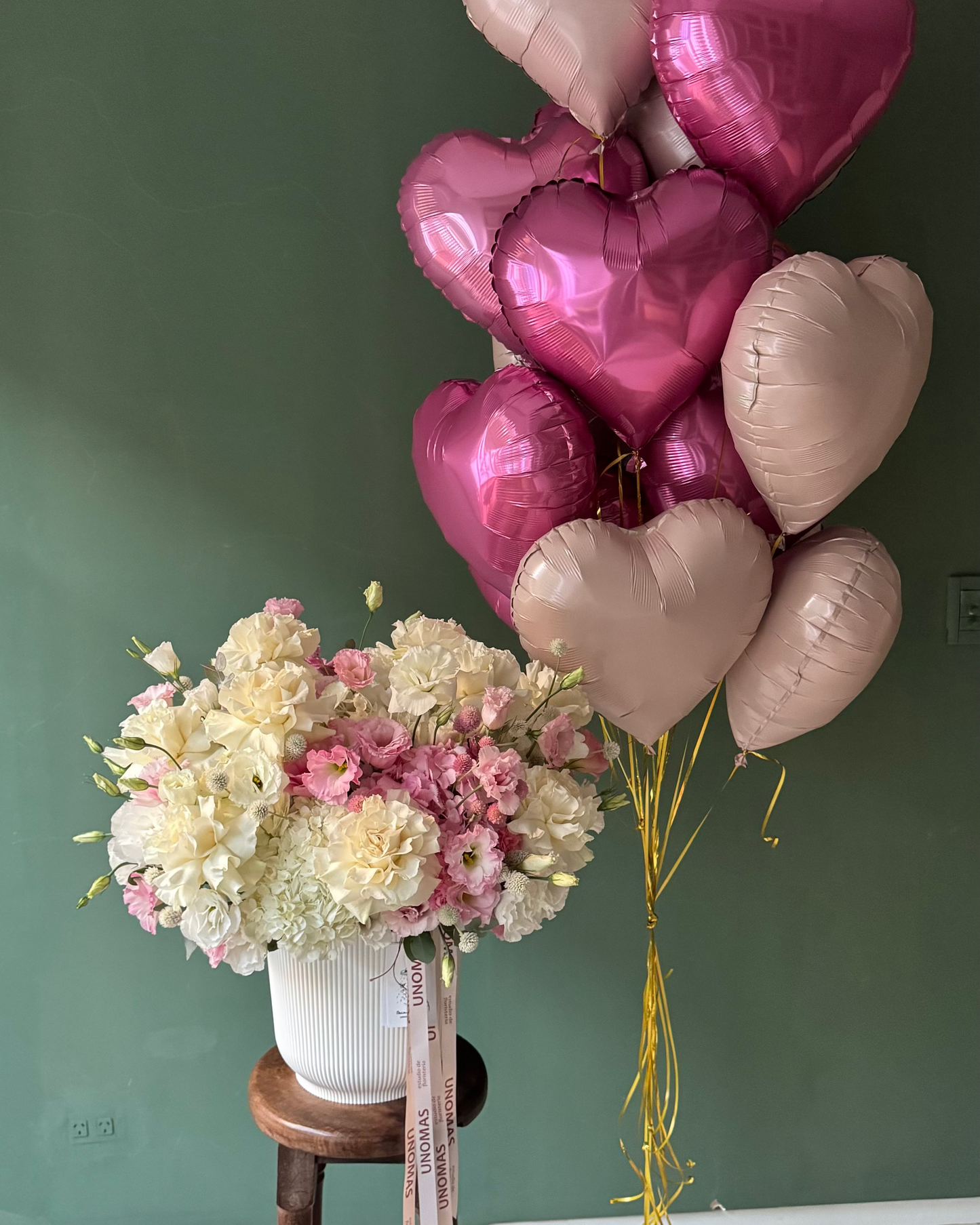 Bouquet of flowers on a stool next to heart-shaped balloons against a green wall.