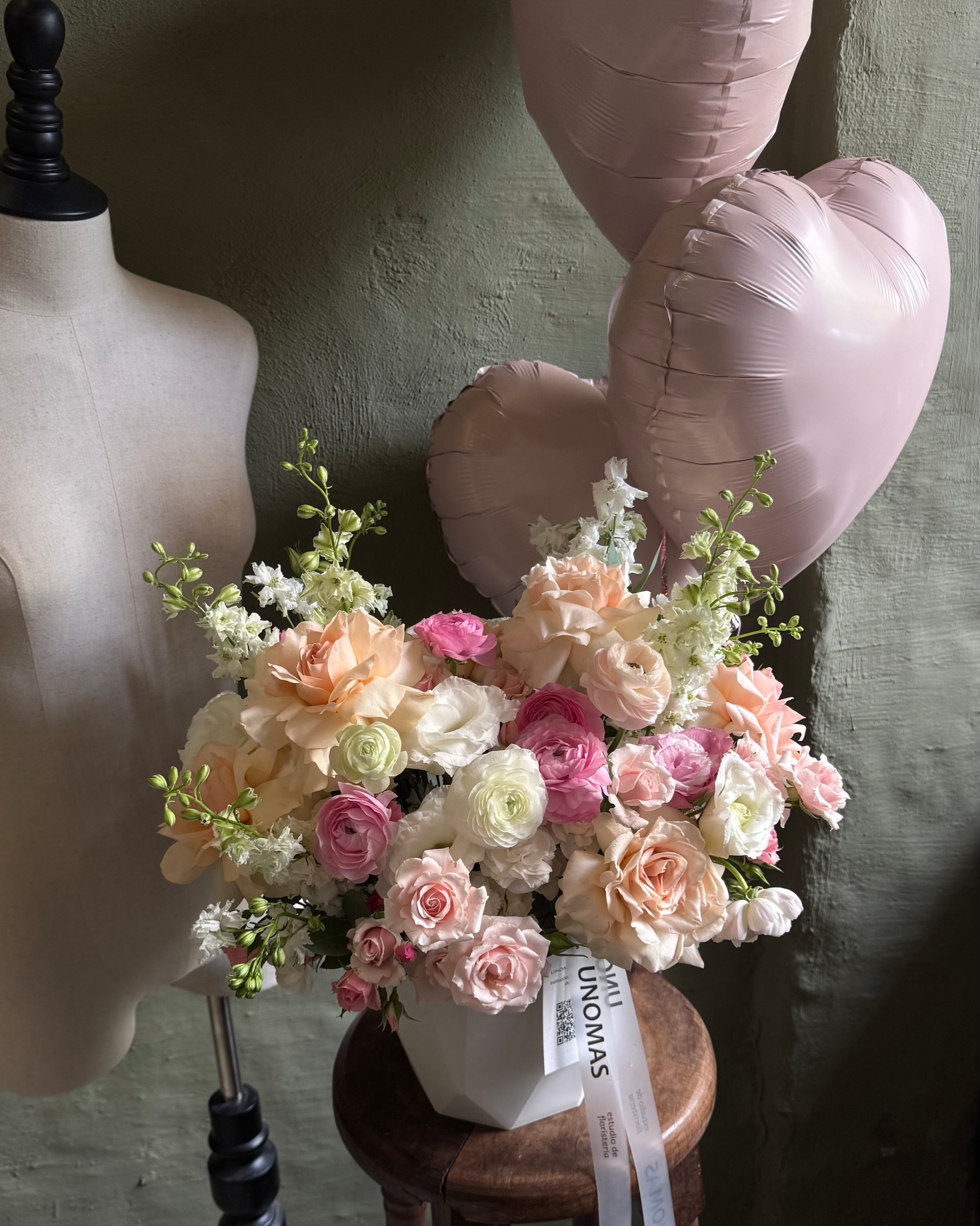 Bouquet of gentle flowers in cachepot on a wooden stool next to a mannequin and pink heart-shaped balloons.