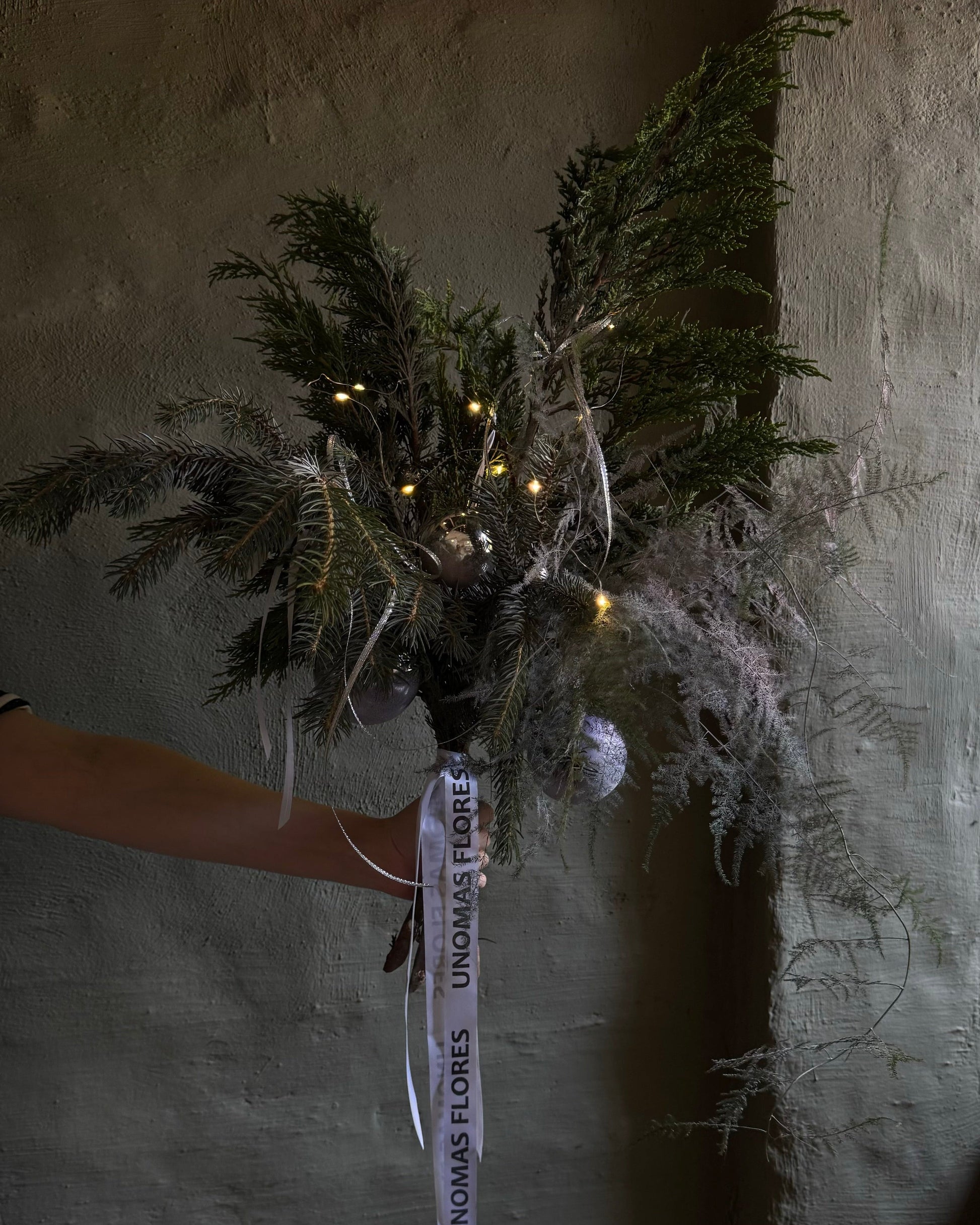 Bouquet of greenery with lights held against a textured wall.