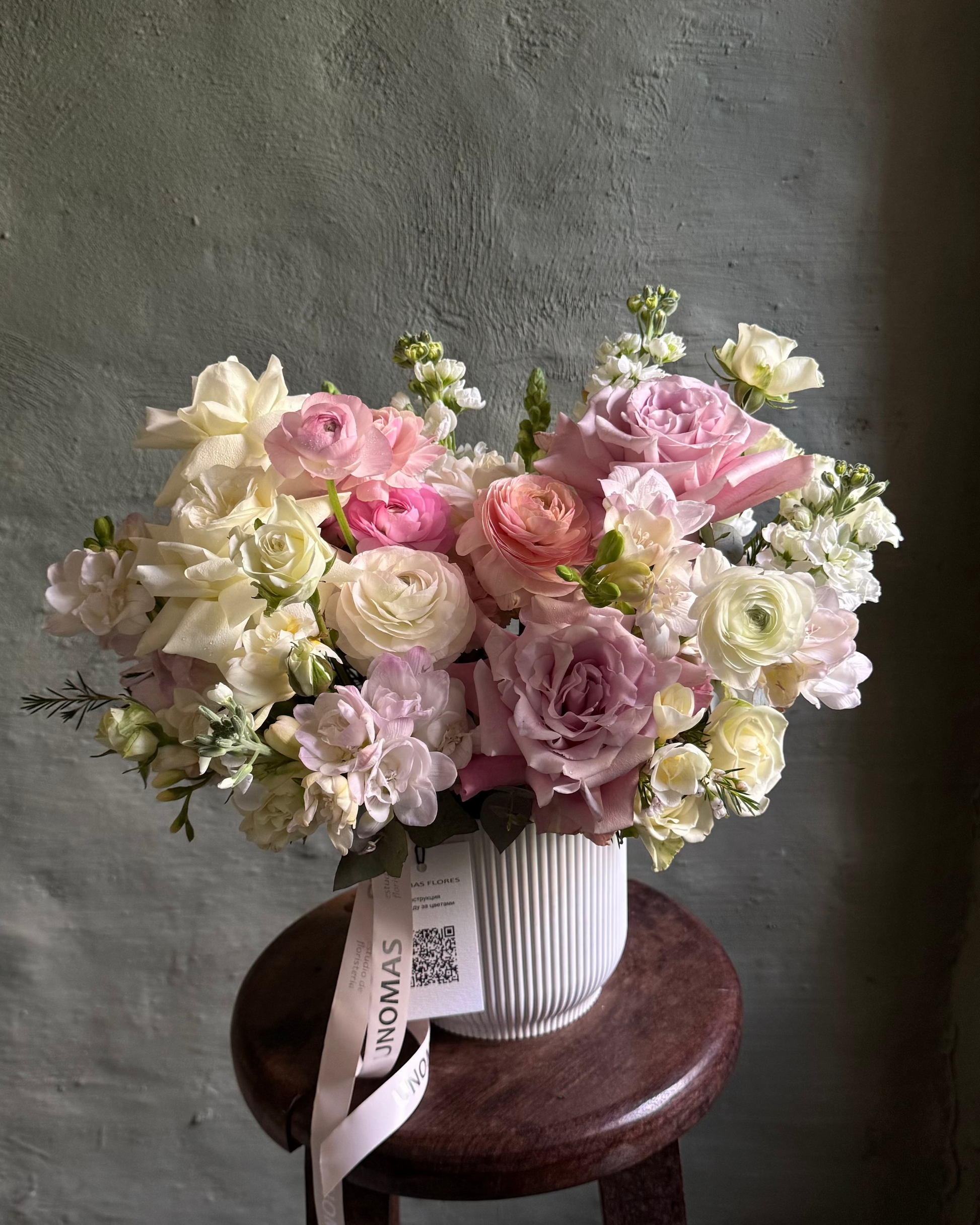 Bouquet of pink and white flowers in a white cachepot on a wooden stool against a gray wall.