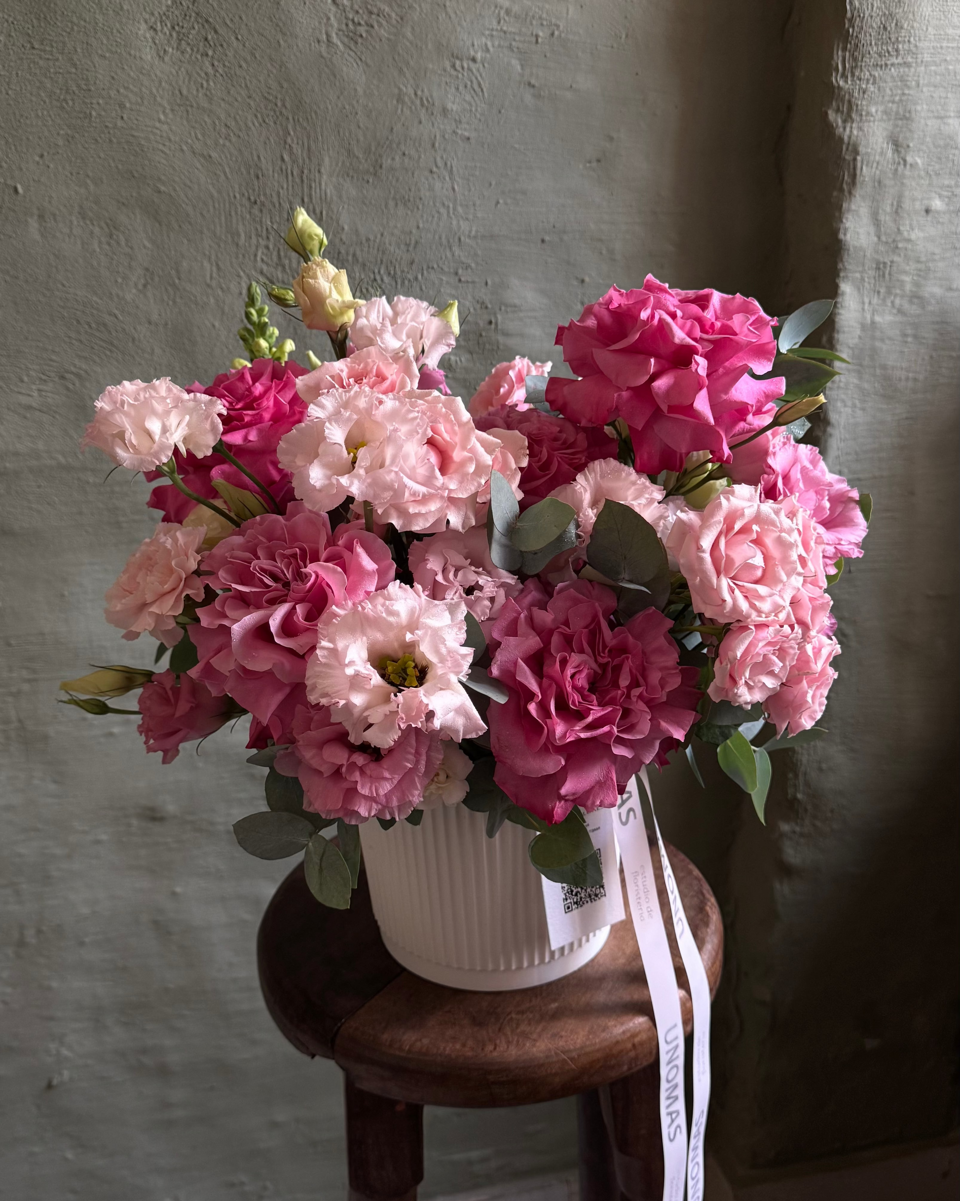 Bouquet of pink and white flowers in a white vase on a wooden stool against a gray wall.