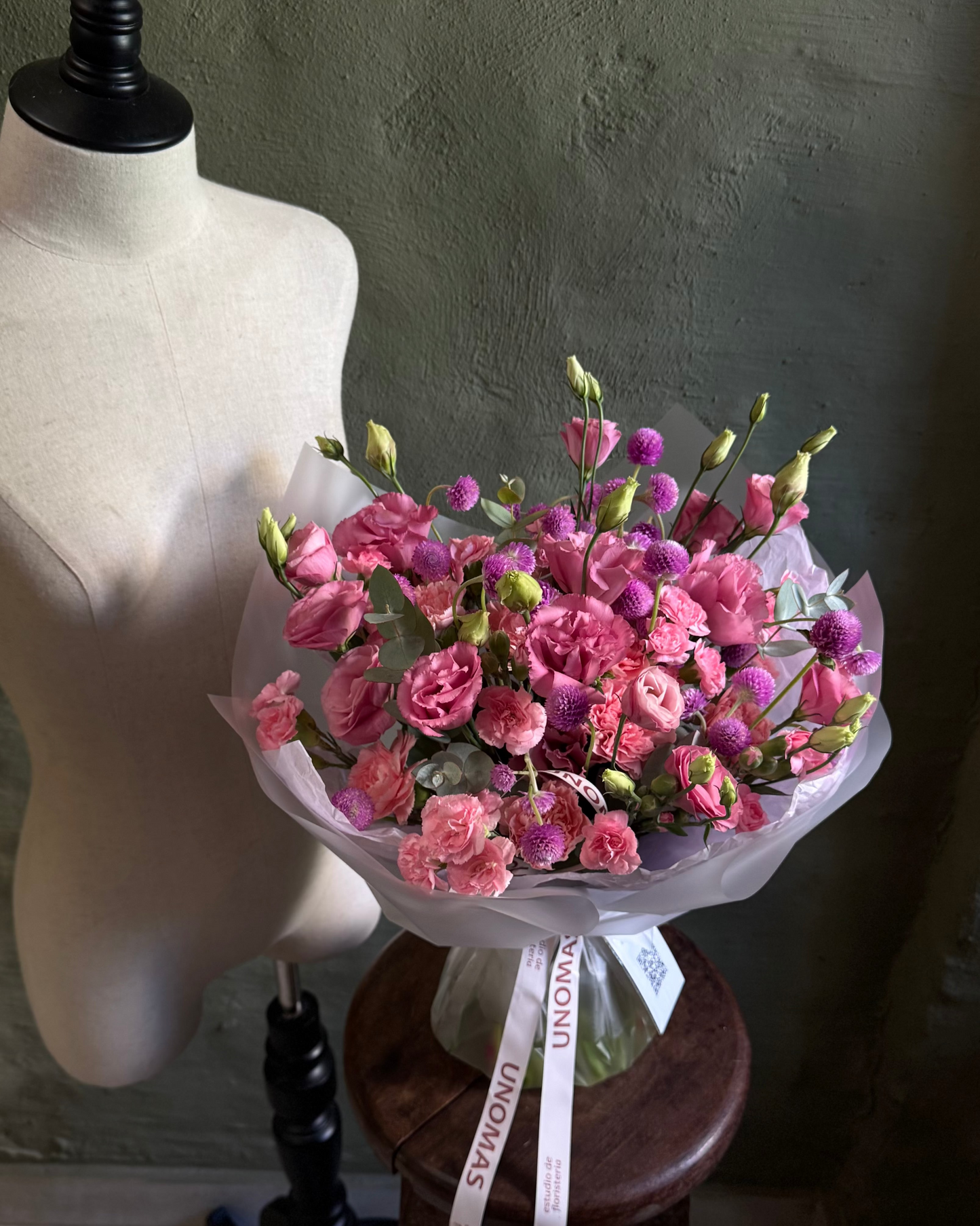 Bouquet of pink flowers on a wooden stool next to a mannequin against a dark wall.