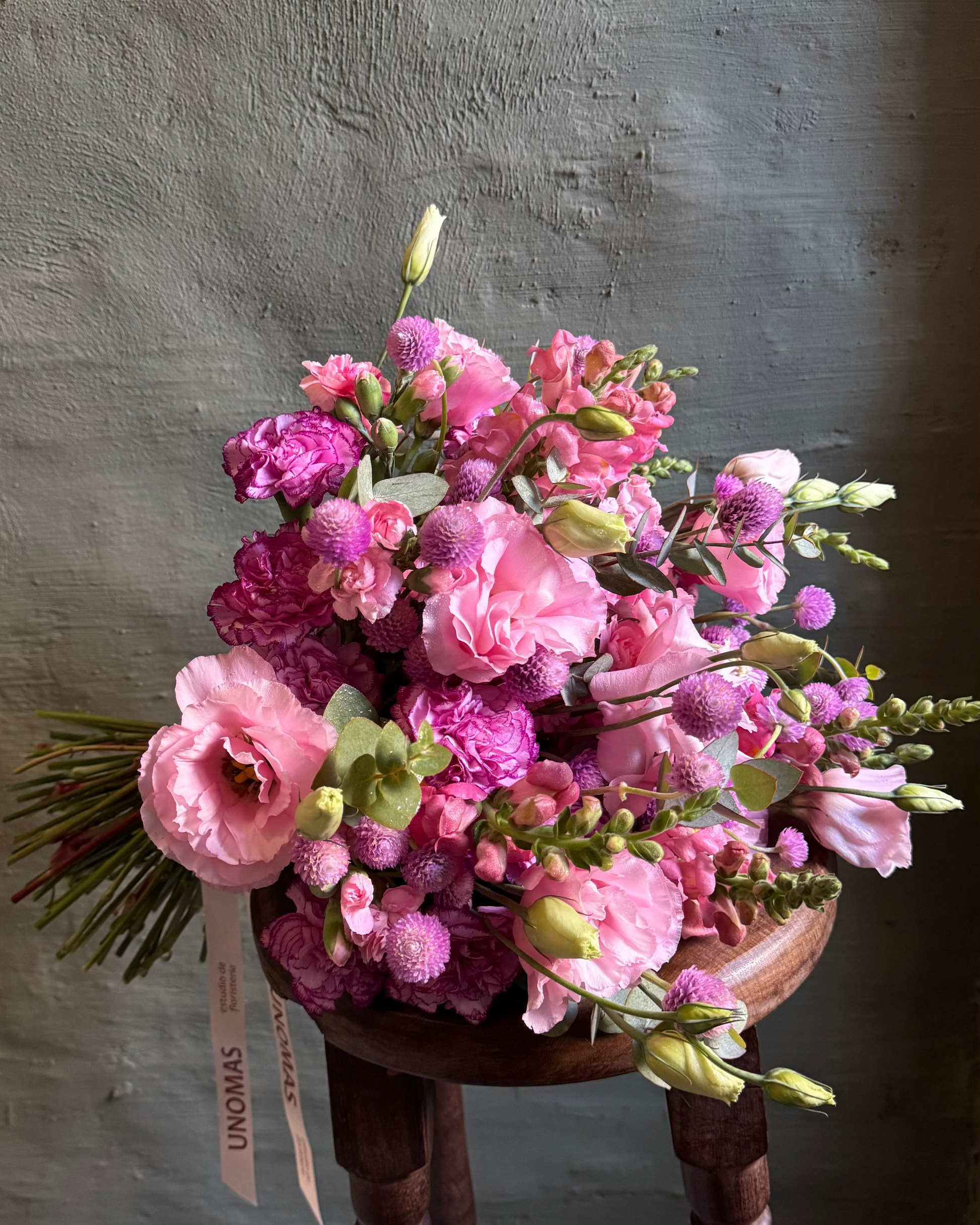 Bouquet of pink flowers on a wooden stool against a textured wall.
