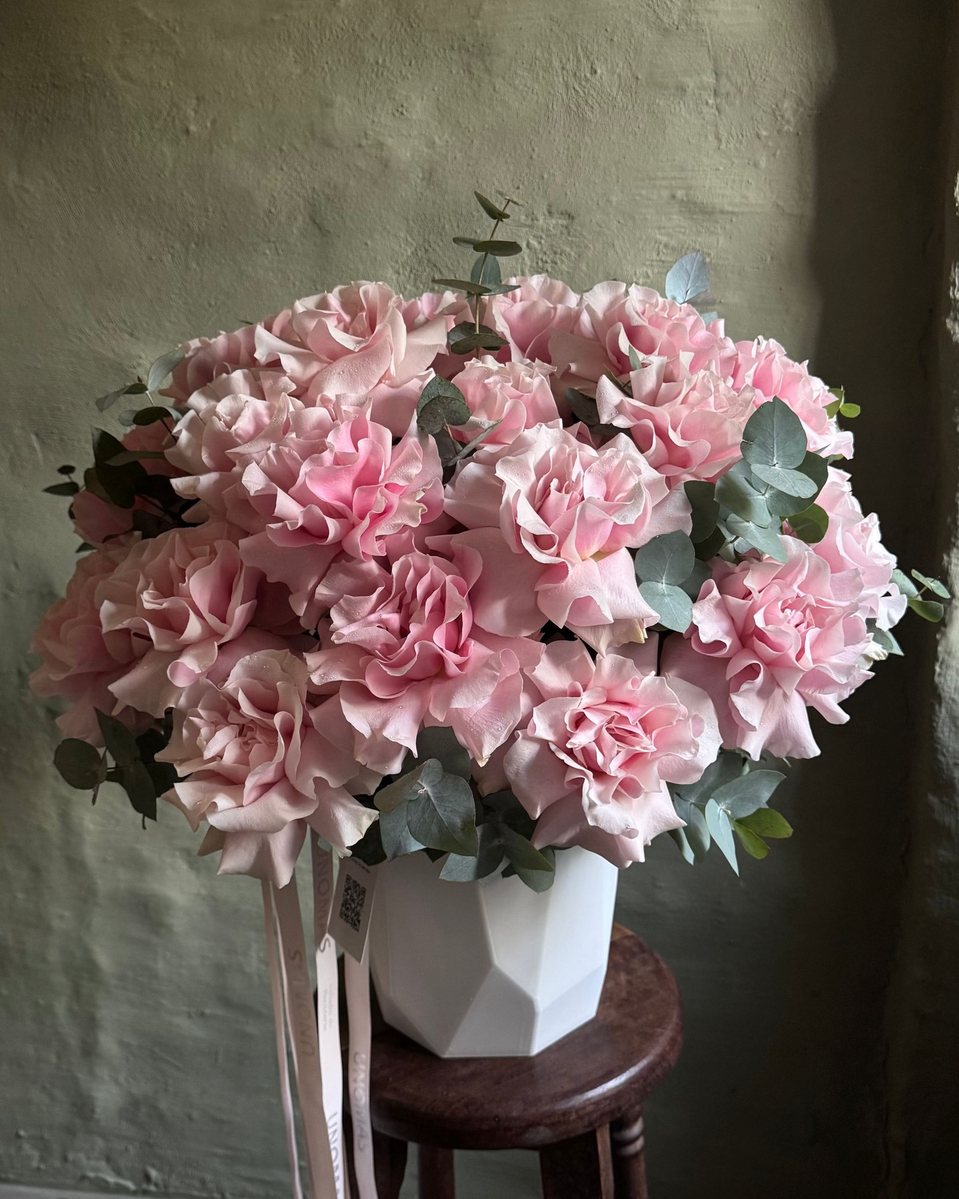 Bouquet of pink flowers in a geometric white vase on a wooden stool against a textured wall.