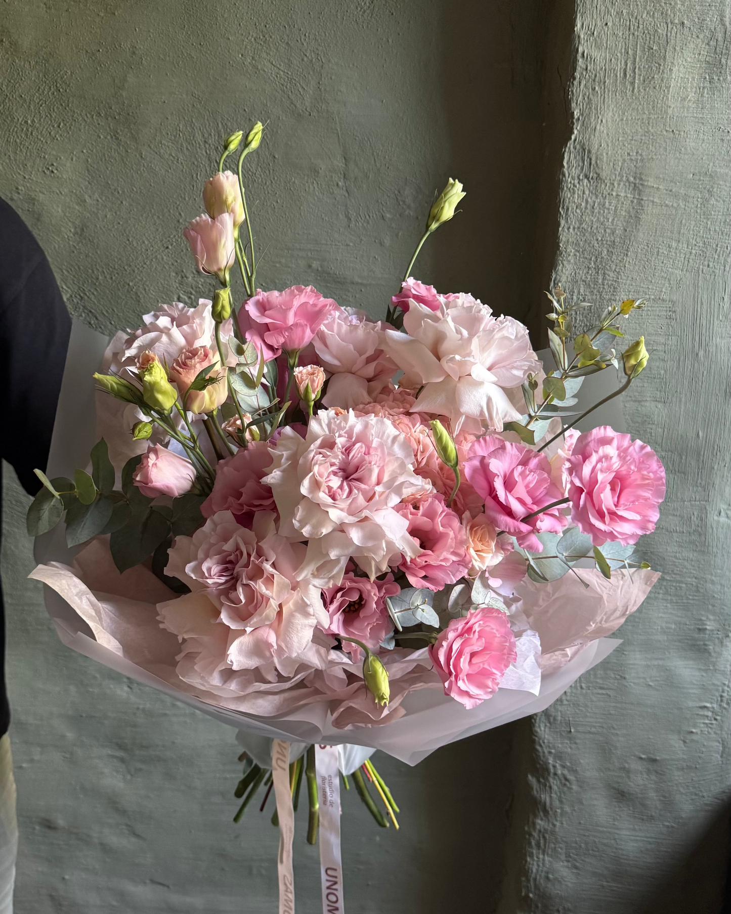 Bouquet of pink flowers wrapped in paper against a textured wall.