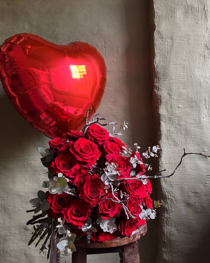 Bouquet of red roses with a heart-shaped balloon on a wooden stool against a textured wall.