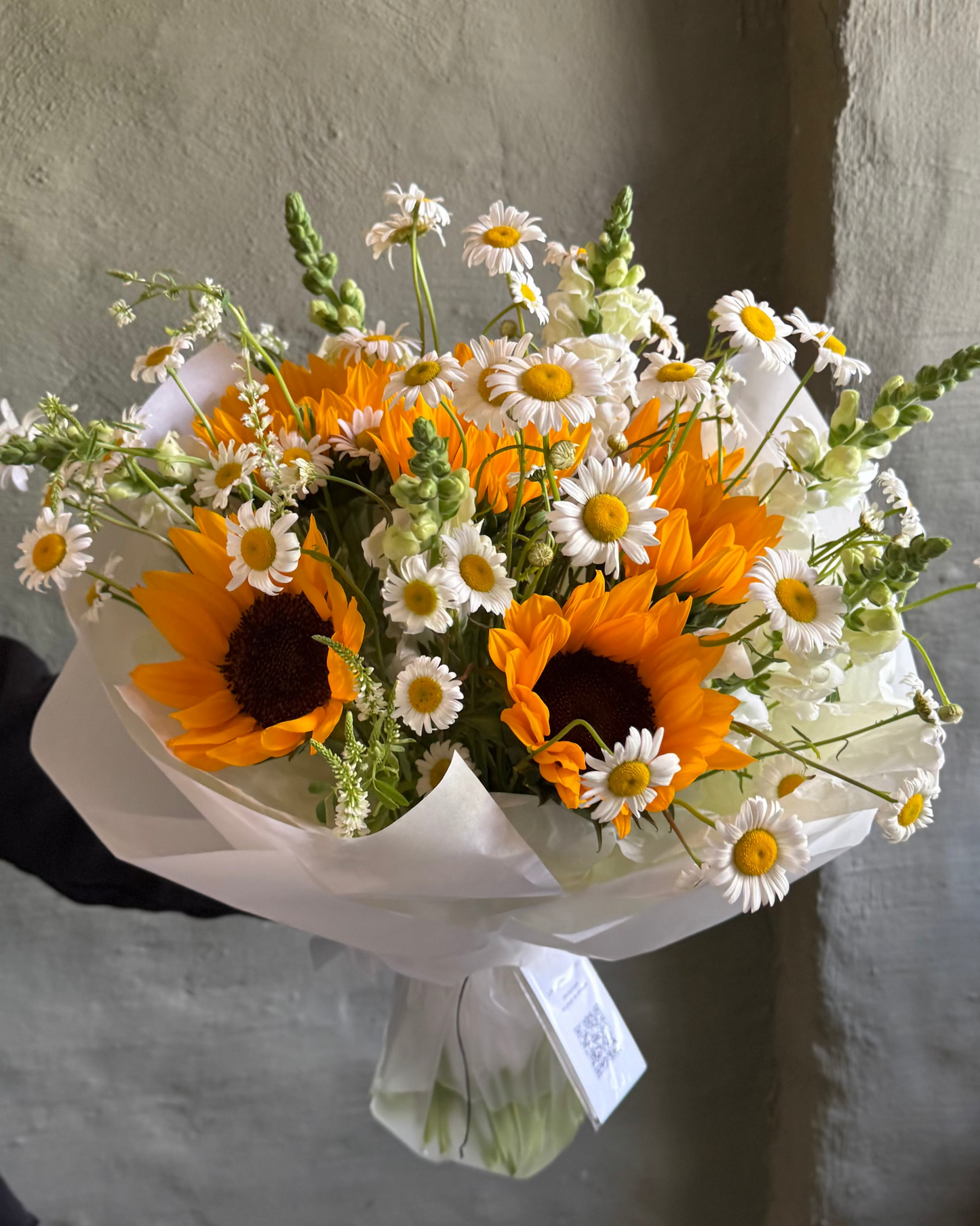 Bouquet of sunflowers and daisies wrapped in white paper against a textured wall.