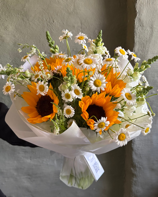 Bouquet of sunflowers and daisies wrapped in white paper against a textured wall.