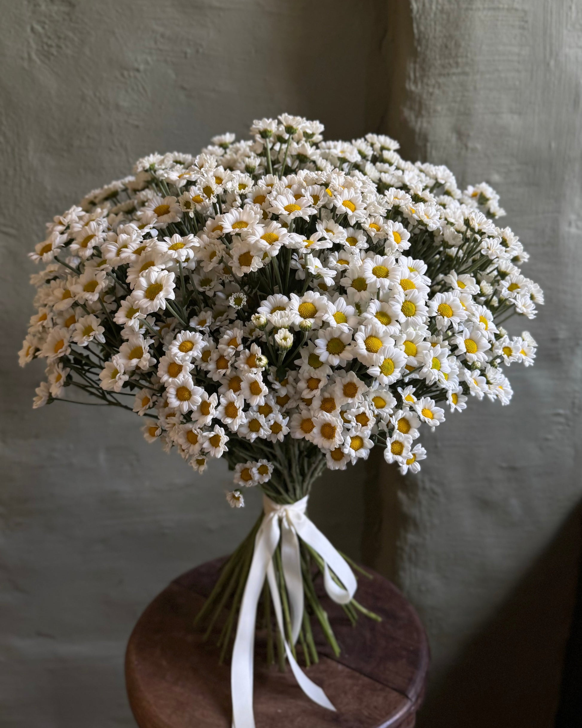 Bouquet of white flowers with green stems on a wooden stool against a textured wall.