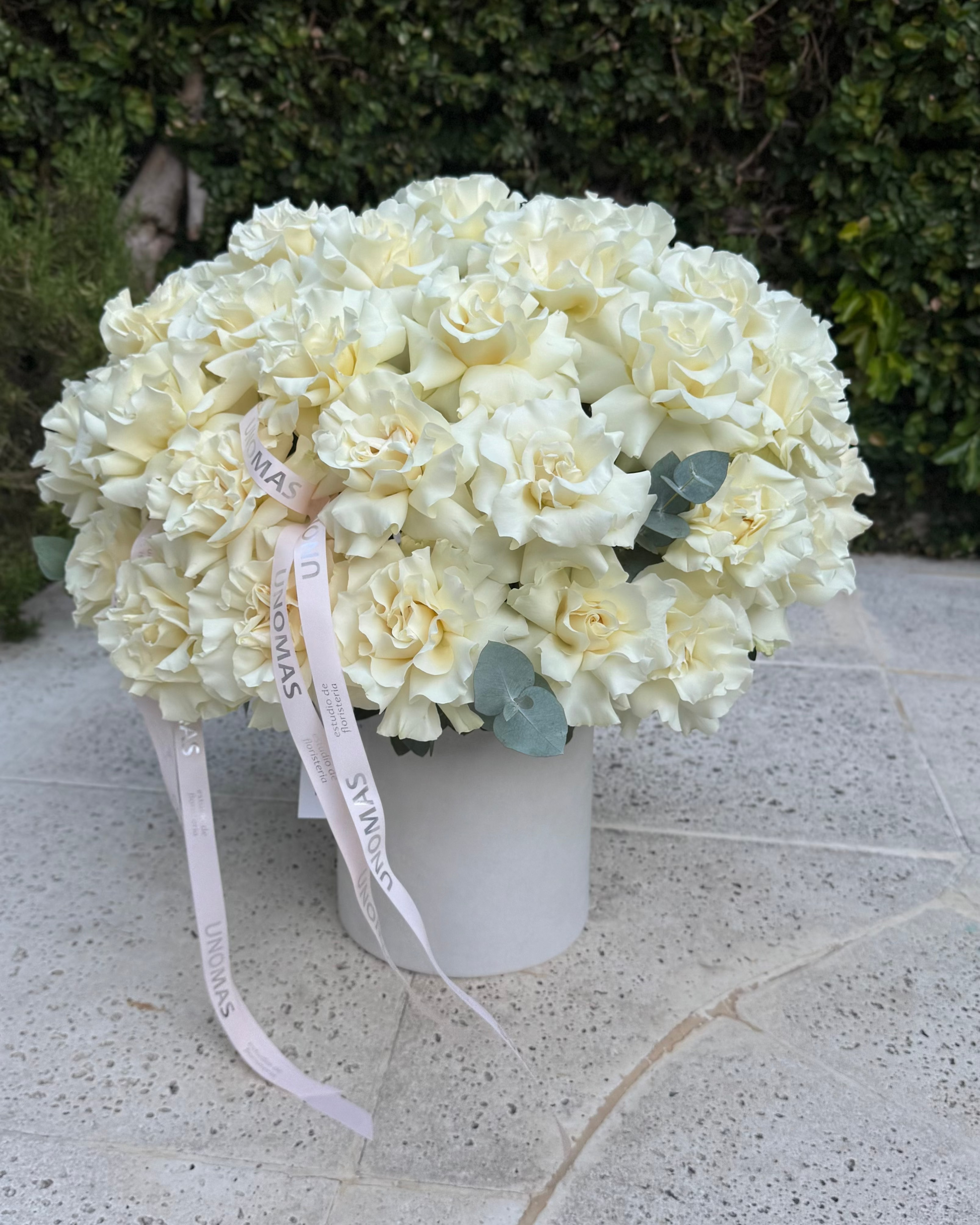 Bouquet of white flowers in a white vase on a stone surface with greenery in the background