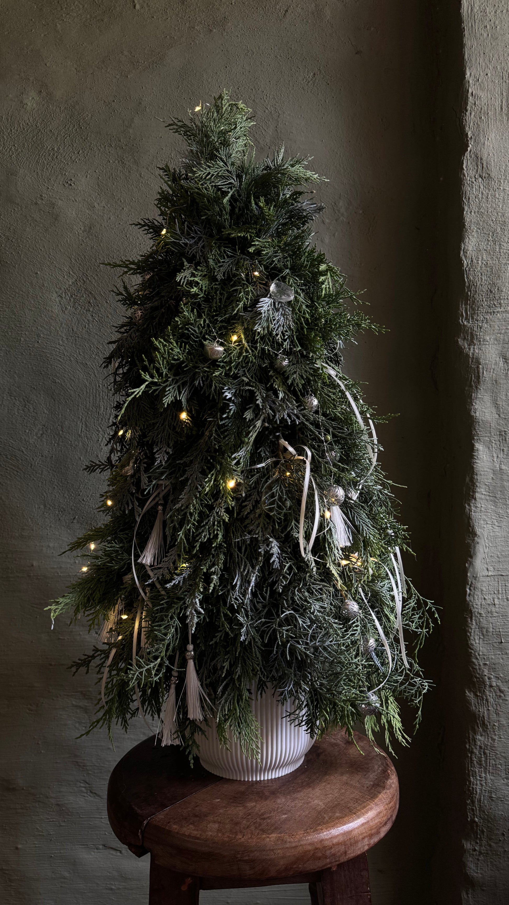 Decorated Christmas tree with lights on a wooden stool against a dark wall