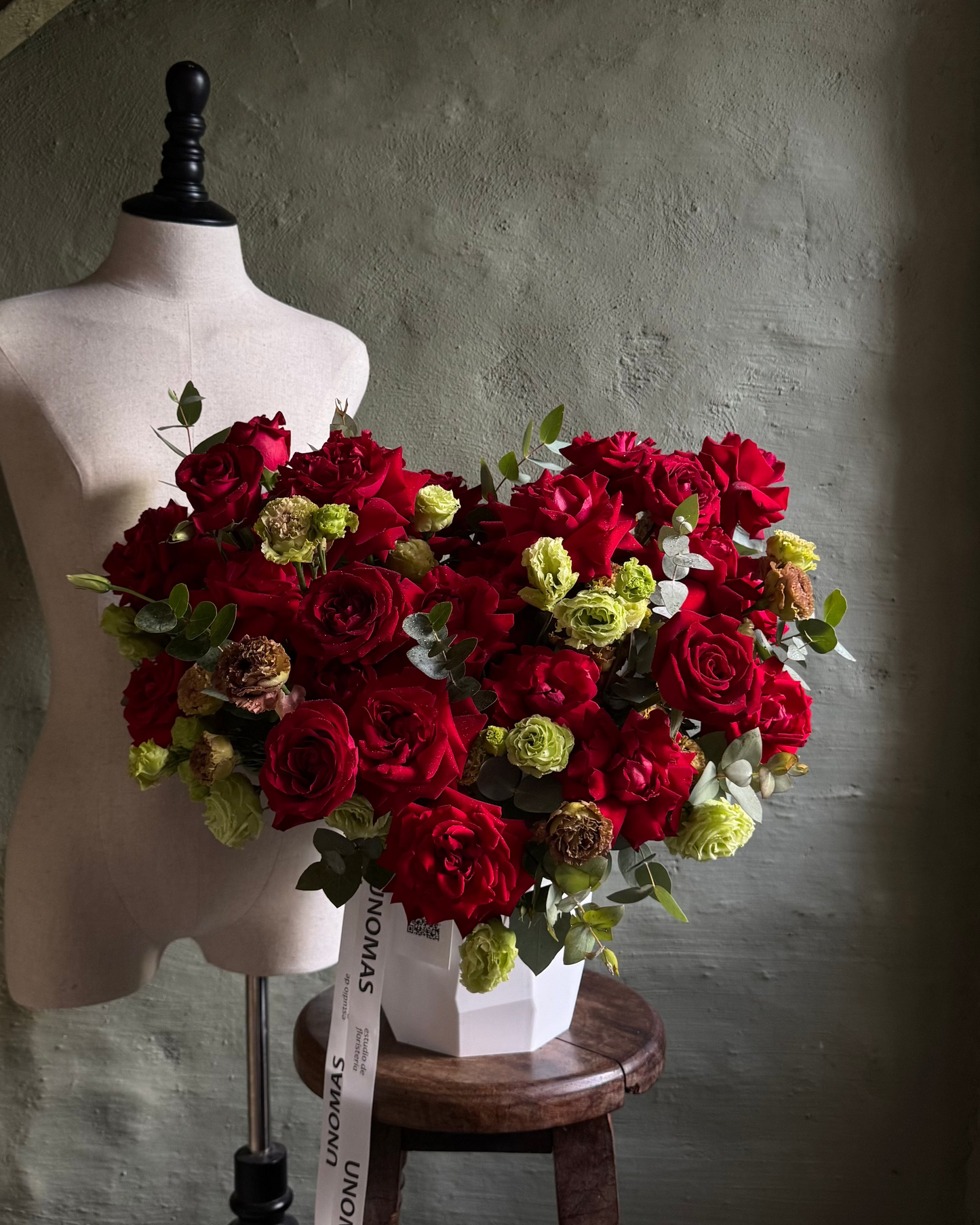 Heart-shaped arrangement of red and green flowers on a wooden stool against a gray wall.