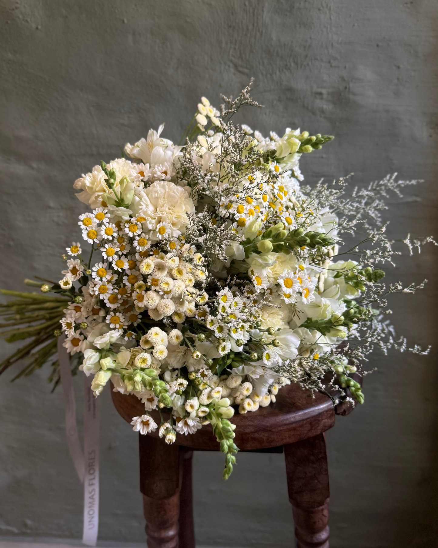 Bouquet of white and yellow flowers on a wooden stool against a gray wall.