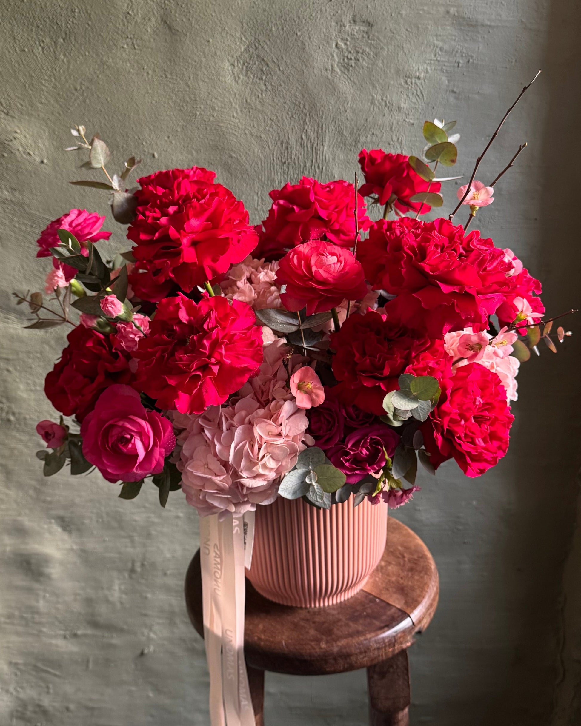 Bouquet of red and pink flowers in a pink vase on a wooden stool against a gray wall.