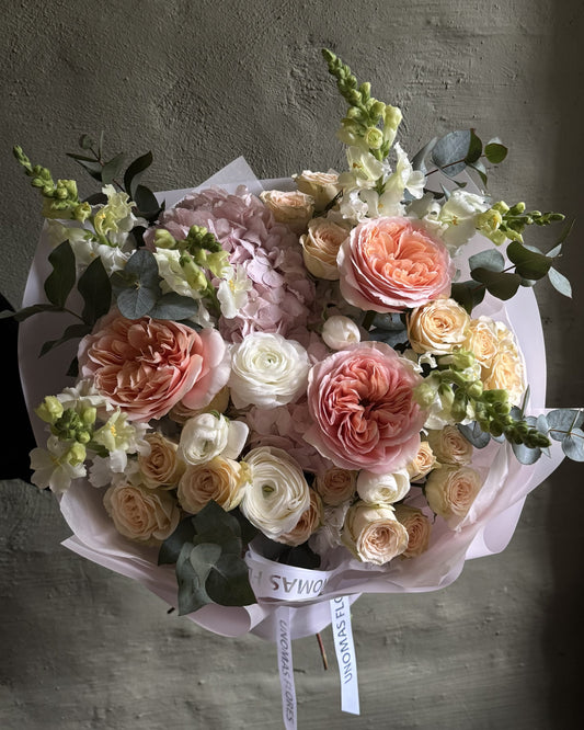 Ramo de rosas durazno y crema, ranúnculos y hortensias rosadas con eucalipto, envuelto en papel rosa claro.