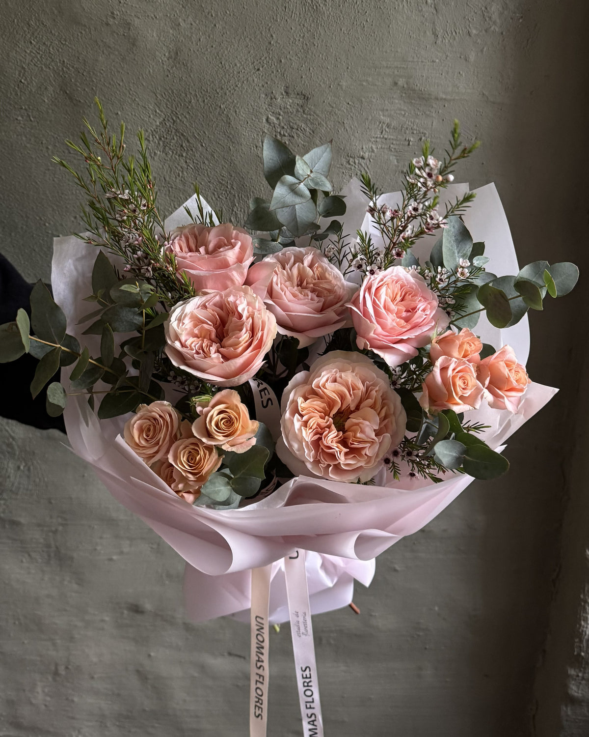 Ramo de rosas en tonos durazno y rosado con eucalipto, envuelto en papel rosa y atado con una cinta.
