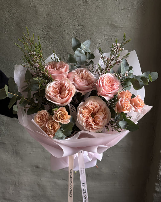 Ramo de rosas en tonos durazno y rosado con eucalipto, envuelto en papel rosa y atado con una cinta.