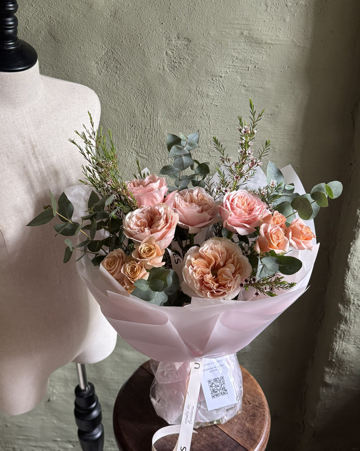 Ramo de rosas durazno y rosadas con eucalipto, apoyado sobre un banquito junto a un maniquí.