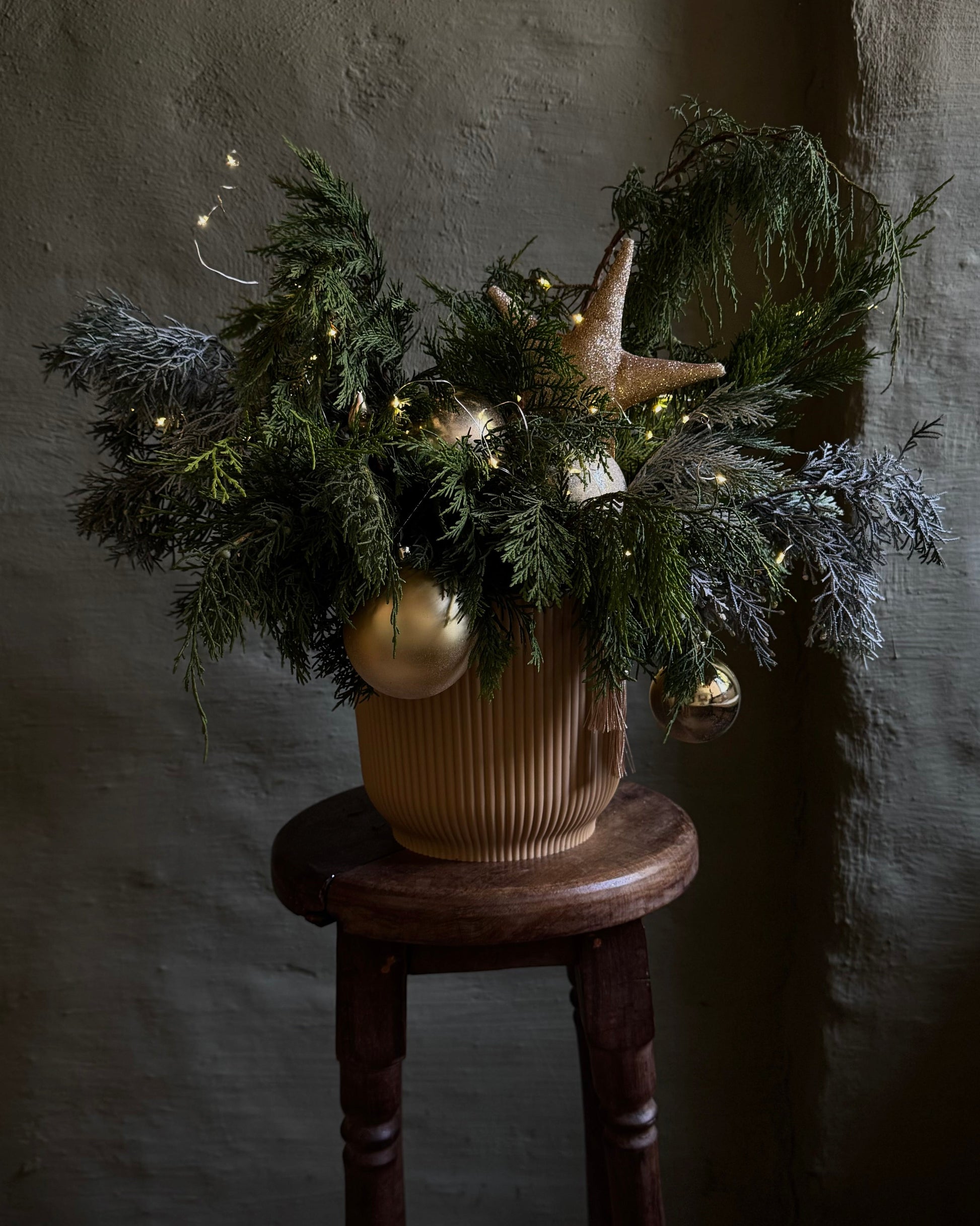 Decorative arrangement of greenery with gold ornaments on a wooden stool against a textured wall.