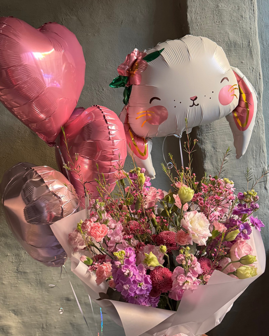 Bouquet of flowers with a rabbit-shaped balloon and heart-shaped balloons on a concrete surface.