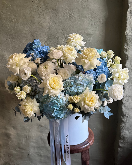 Bouquet of blue and white flowers in a white vase on a wooden stool against a gray wall.