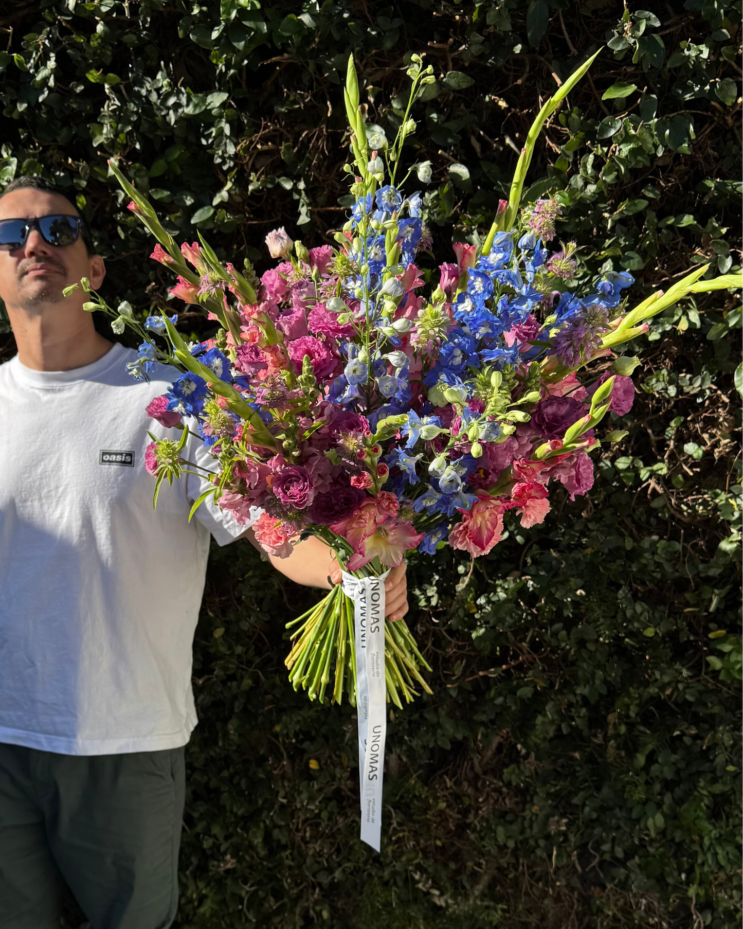 Man holding a bouquet of flowers against a green hedge.