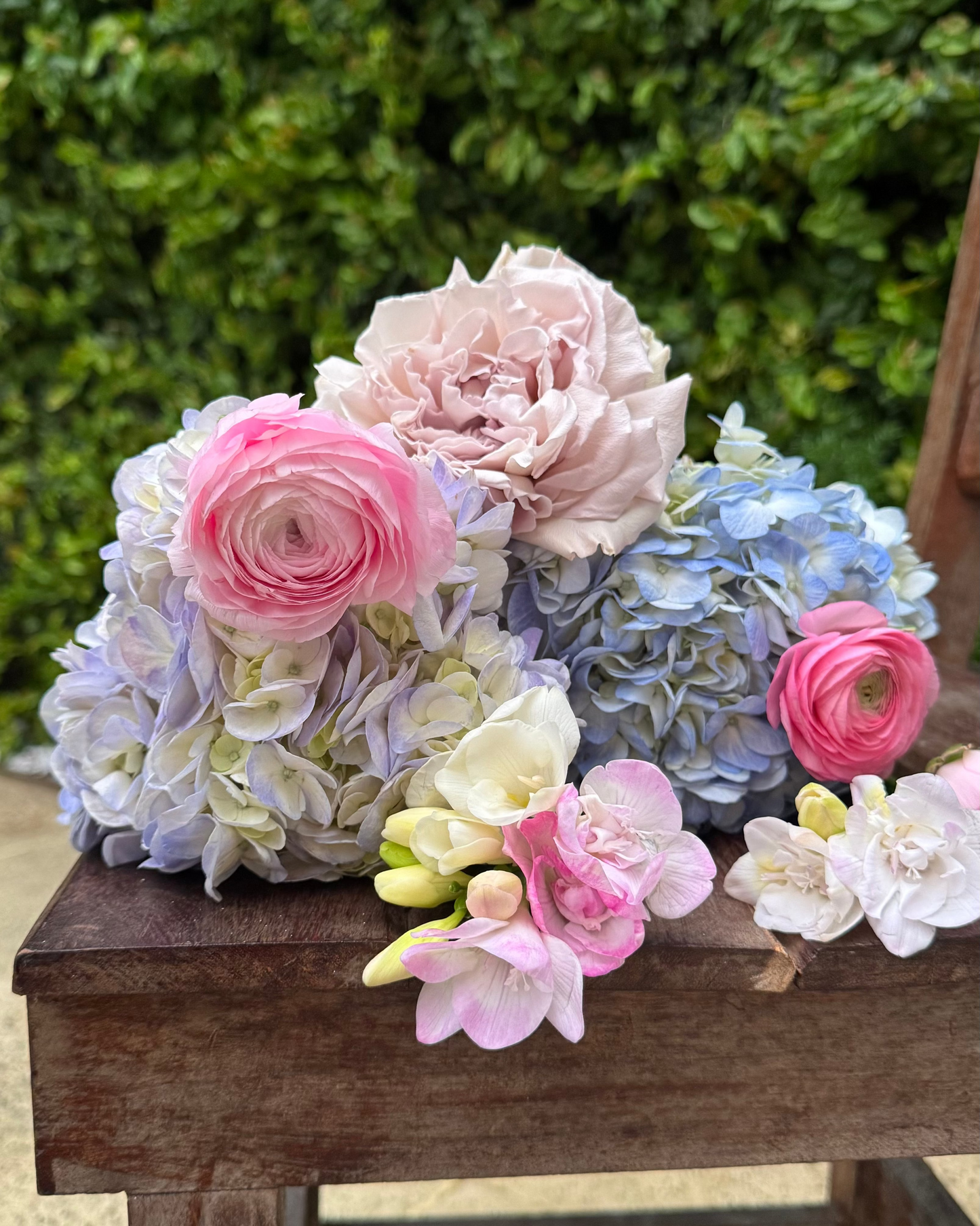 Colorful flower arrangement on a wooden bench with a green bush background