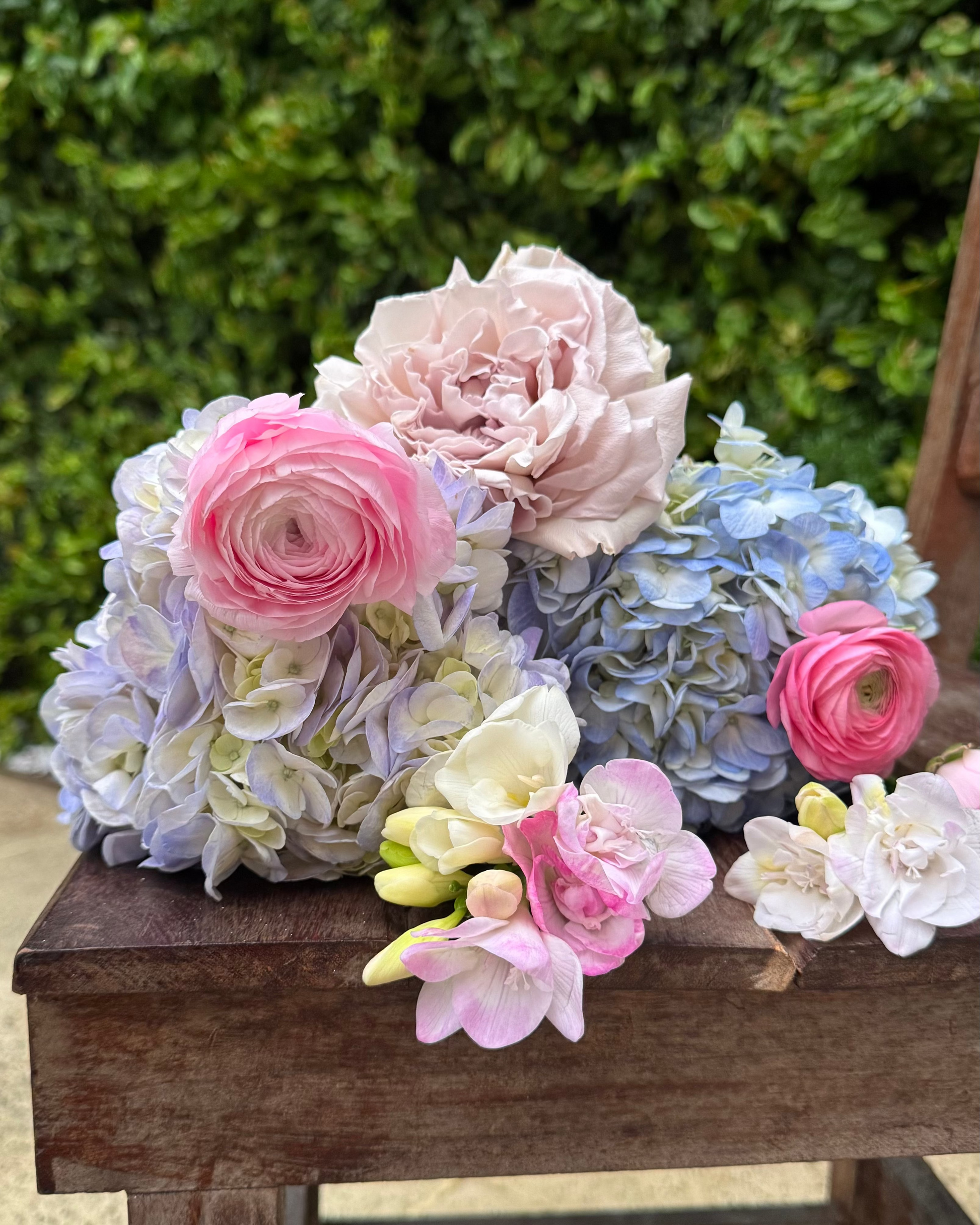 Colorful flower arrangement on a wooden bench with a green bush background