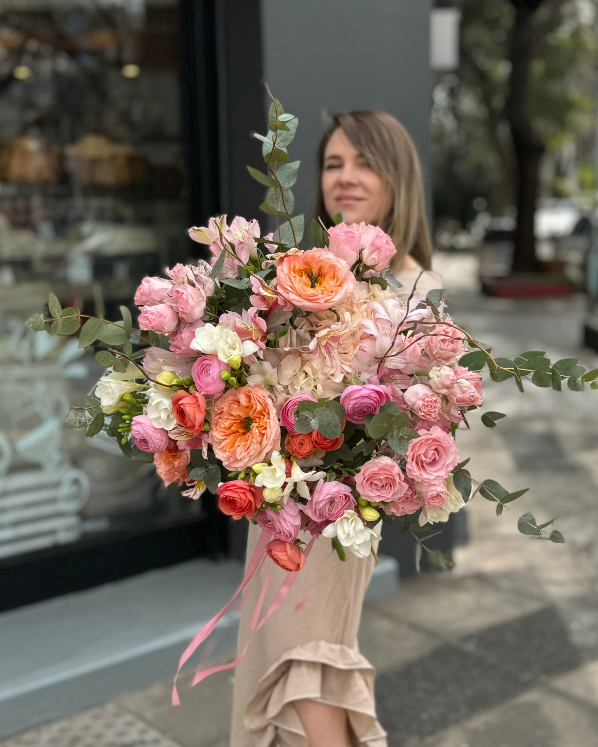 Ramo de rosas color durazno, hortensias rosadas y ranúnculos con follaje, sostenido por una mujer frente a la florería.