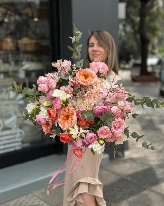 Ramo de rosas color durazno, hortensias rosadas y ranúnculos con follaje, sostenido por una mujer frente a la florería.