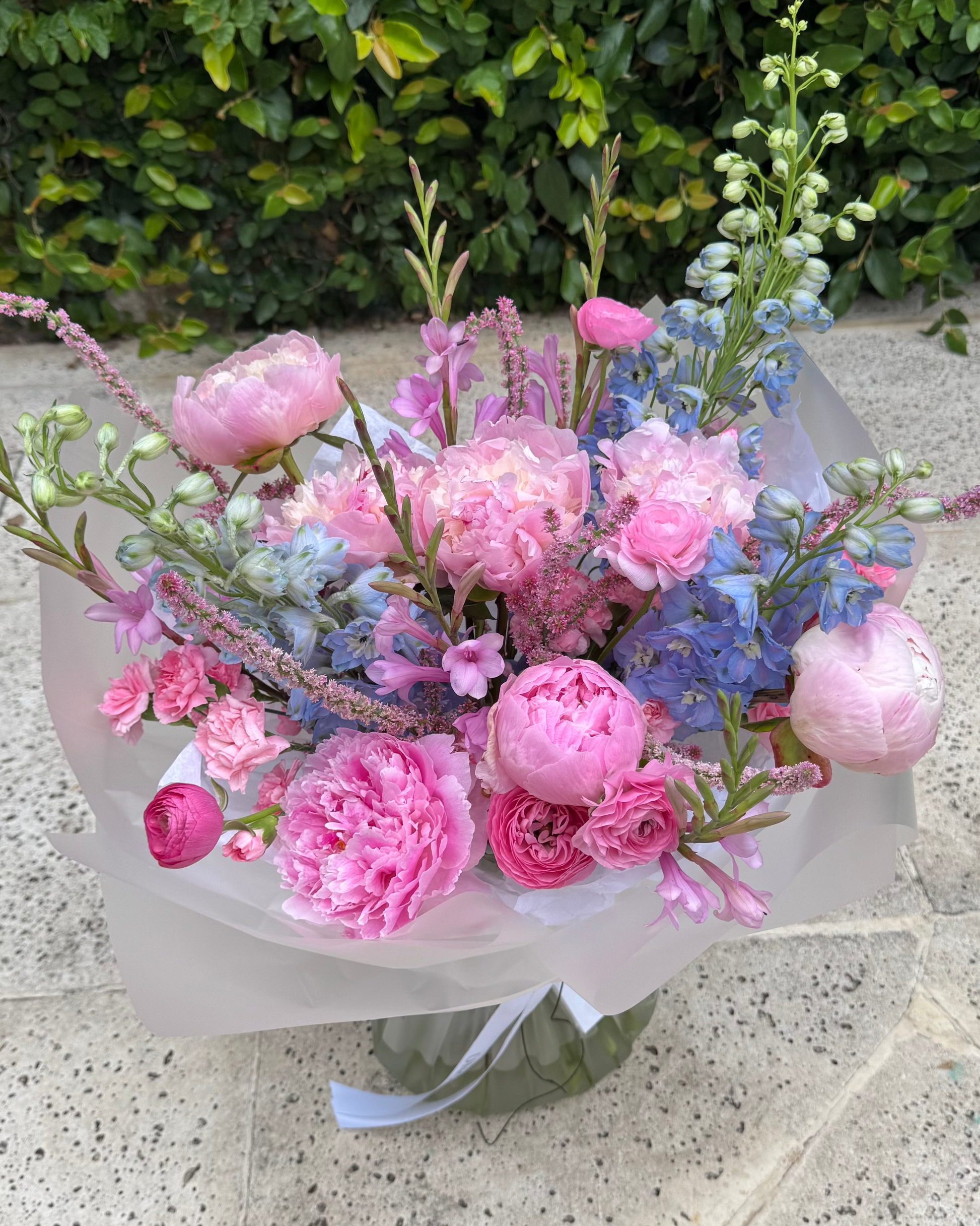 Bouquet of pink and blue flowers in a clear vase on a stone surface with greenery in the background