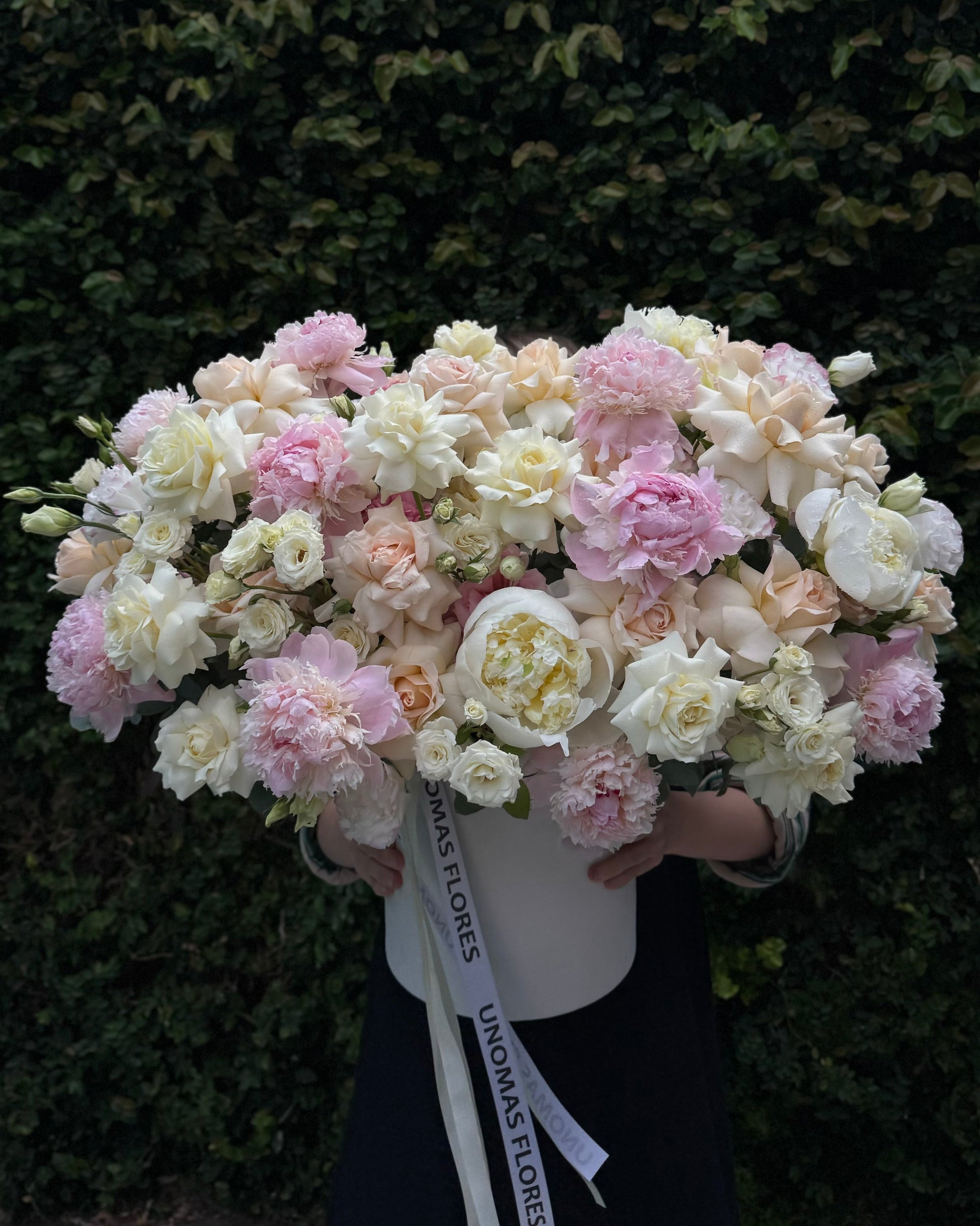 Person holding a large bouquet of pink and white flowers against a green hedge.