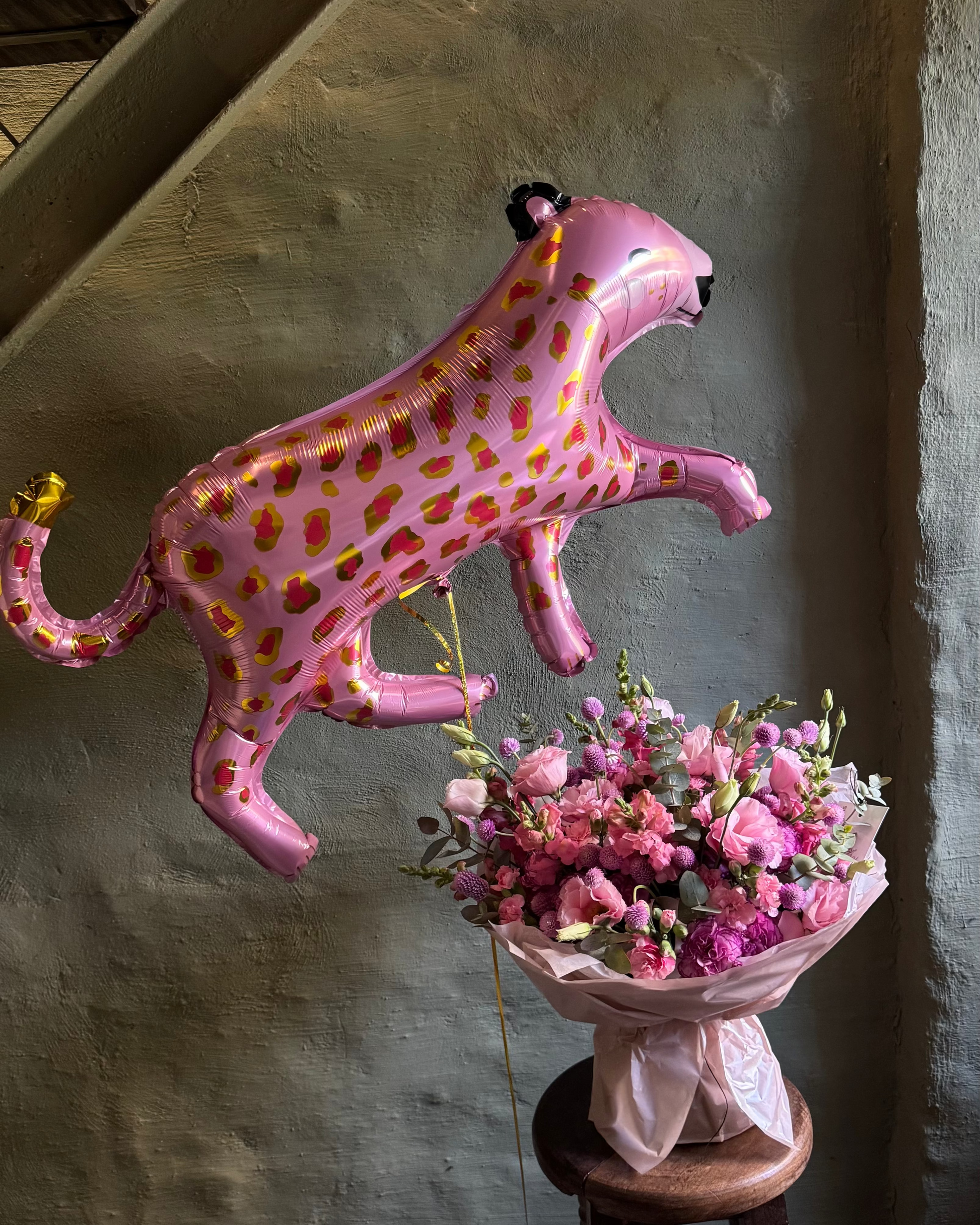 Pink panther balloon and bouquet of flowers on a wooden stool against a textured wall.